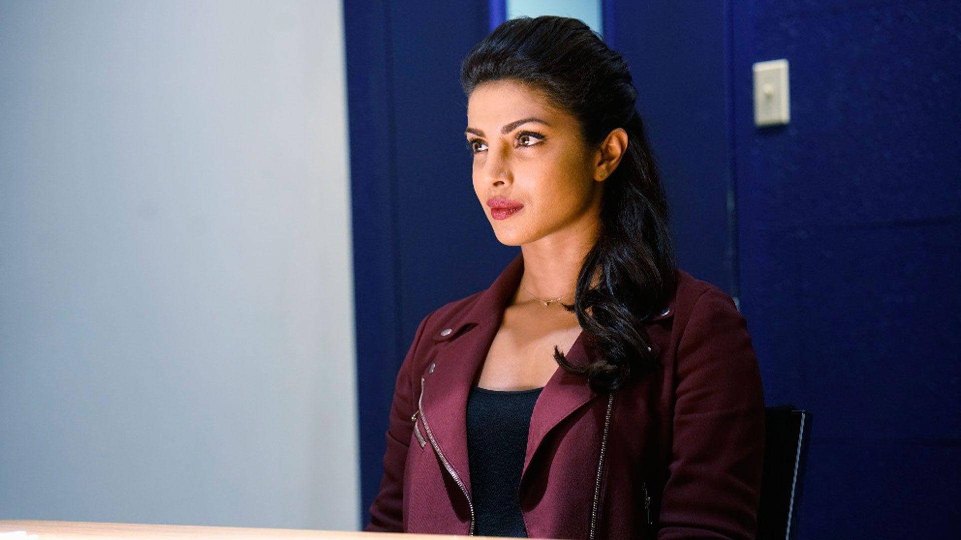 《Quantico》FBI探員艾莉克斯·帕里什 壁紙 — 一位身穿栗色外套的女性坐在桌子旁，面對藍色牆壁，表情嚴肅。