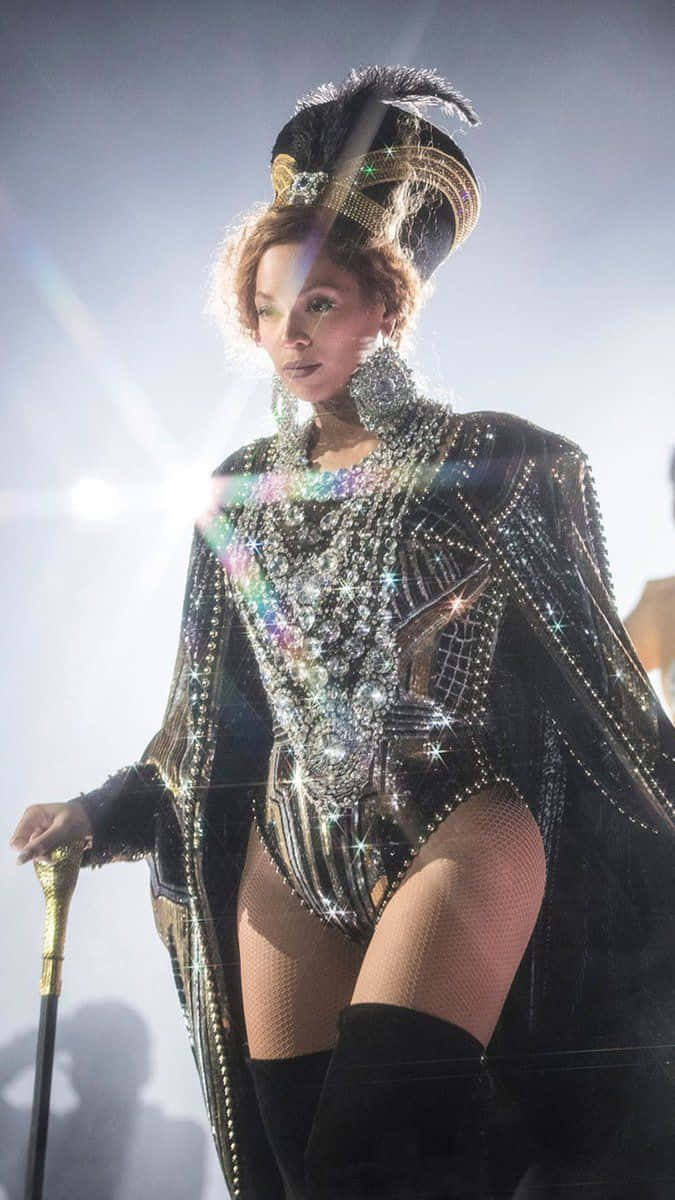 Queen Bey擺姿 壁紙 — Beyoncé 身穿閃耀的黑金珠寶緊身連衣褲，頭戴羽毛頭飾，手持手杖，被明亮的舞台燈光照亮。
