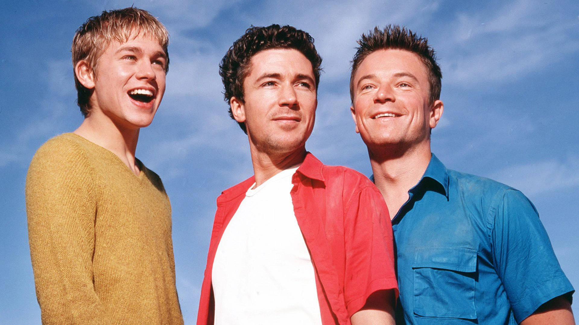 《Queer As Folk》微笑三人組 壁紙 — 三位微笑的年輕男子穿著色彩繽紛的襯衫，站在明亮的藍天白雲下。
