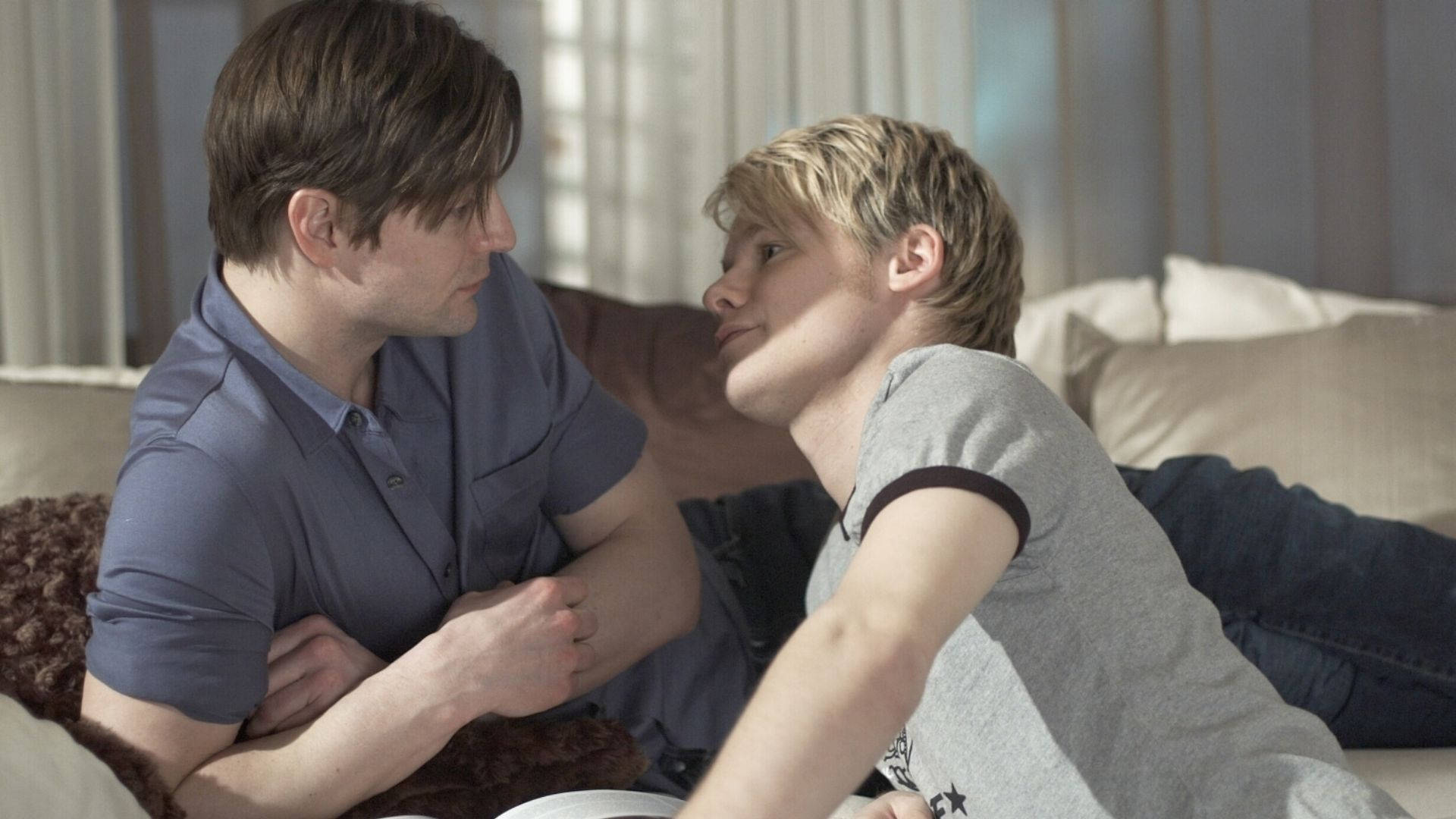 《Queer As Folk》甜蜜凝視 壁紙 — 兩個男人在燈光柔和嘅客廳入面，親密咁坐喺梳化上分享一個親密時刻。