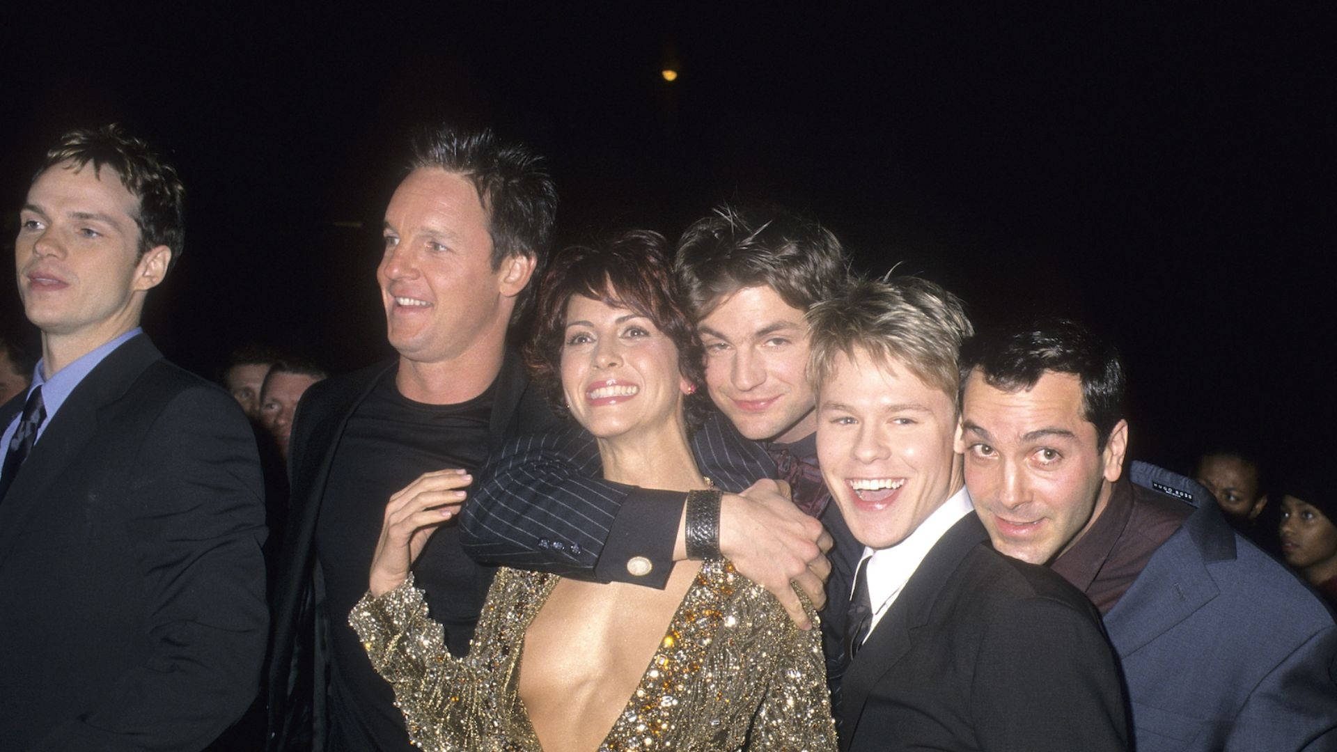 《Queer As Folk》首映夜 壁紙 — 六位微笑的名人在夜間活動中於人造燈光下緊密地一起擺姿勢。