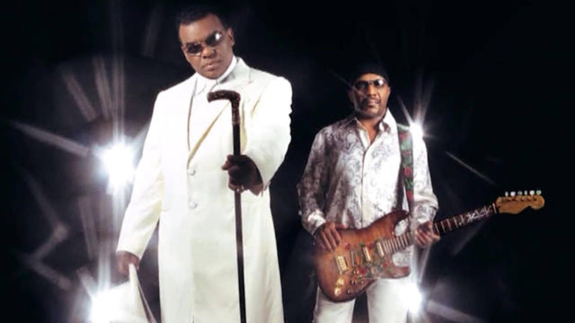 R&B 傳奇樂團 Isley Brothers 與主唱 Ronald Isley 壁紙 — 兩位時尚男士戴著太陽眼鏡，一位身穿白色西裝手持拐杖，另一位拿著結他，在黑色背景下被明亮的舞台燈光照亮。