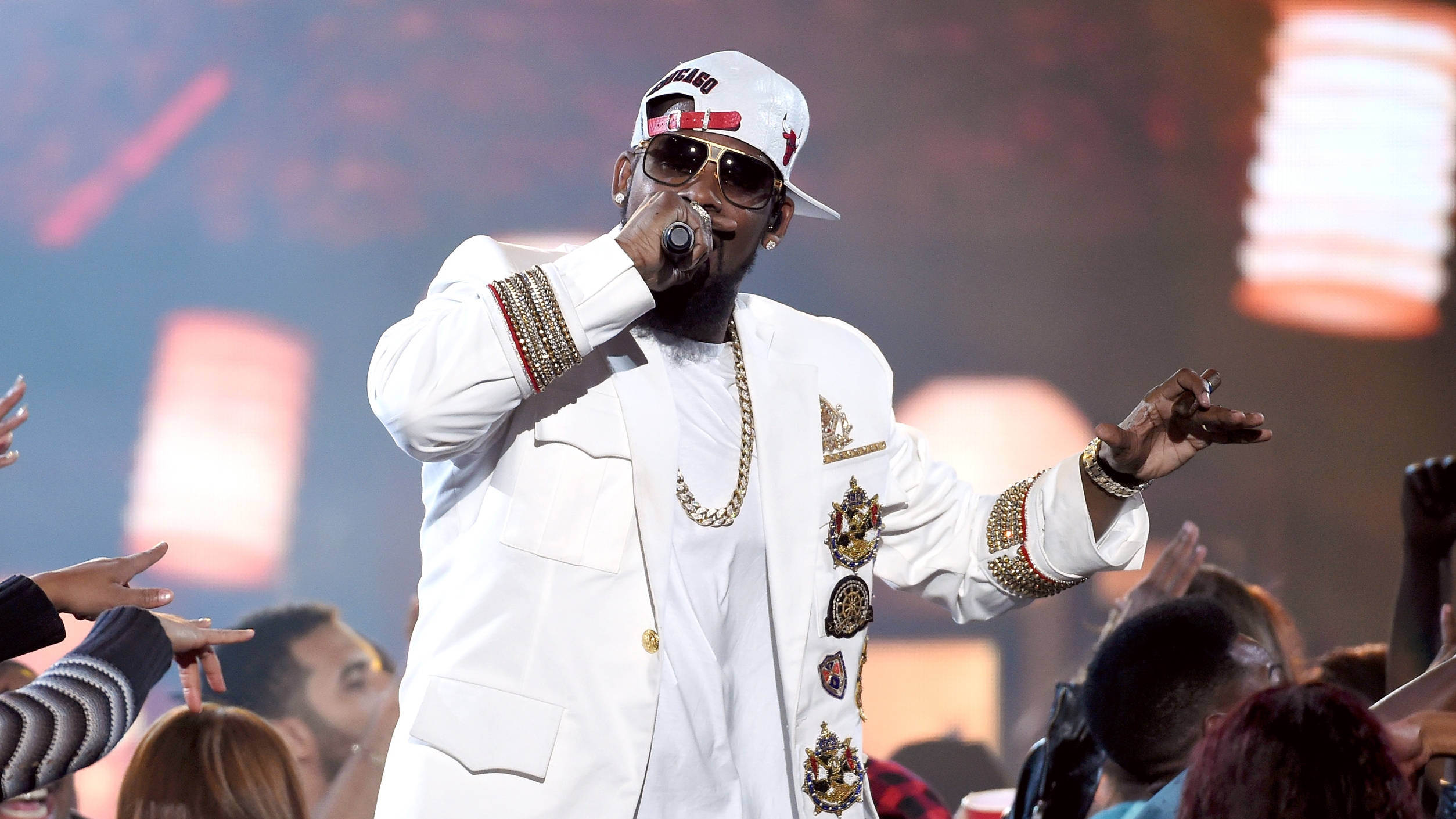 R. Kelly 舞台演唱 壁紙 — 一位身穿白色服裝和太陽眼鏡的說唱歌手在舞台上表演，人群在五彩繽紛的舞台燈光下向他伸出手。