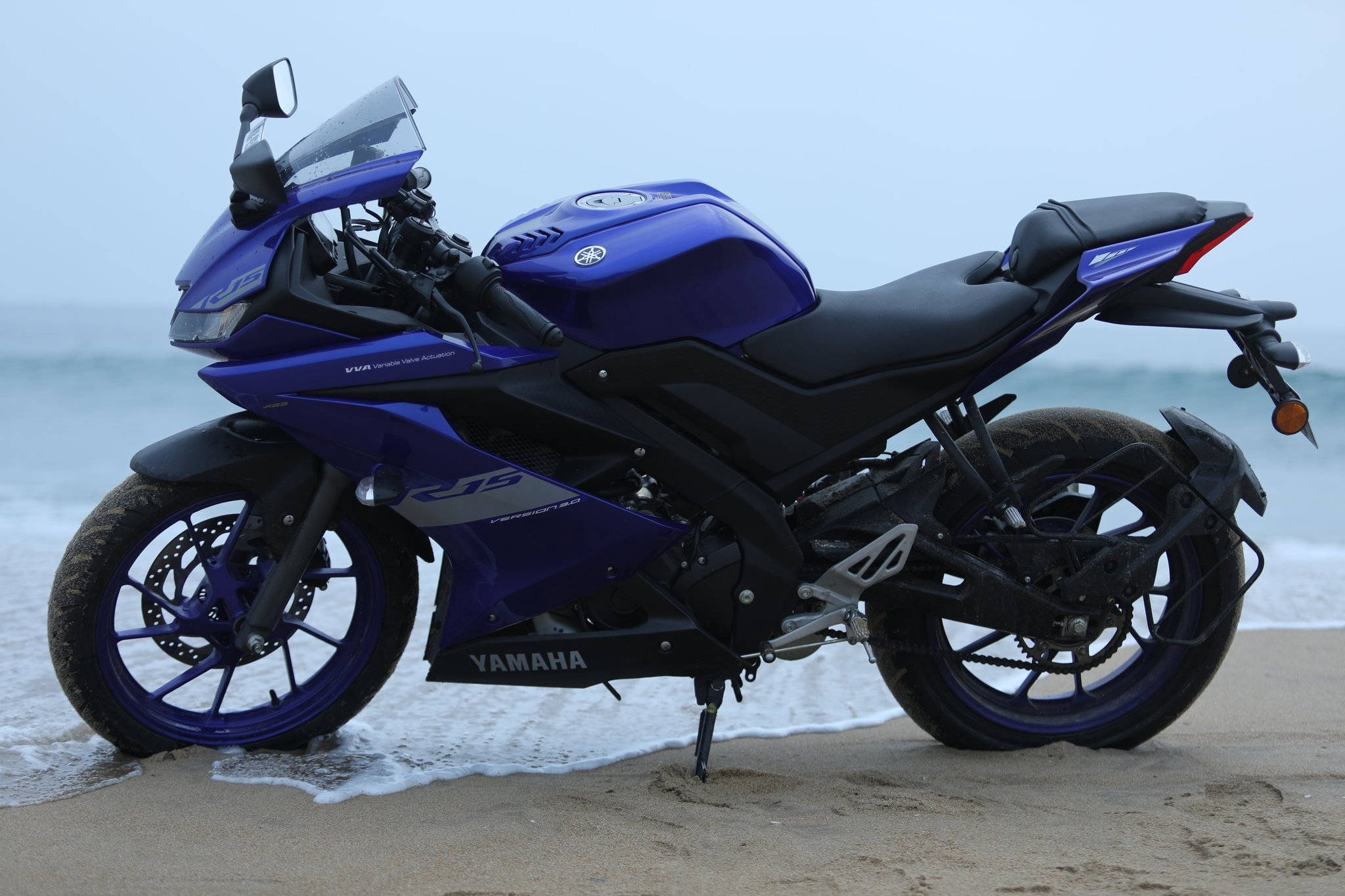 R15 V3 海灘 壁紙 — 一架充滿活力的藍色Yamaha R15運動電單車停在濕沙上，海浪在陰天輕拍其輪胎。