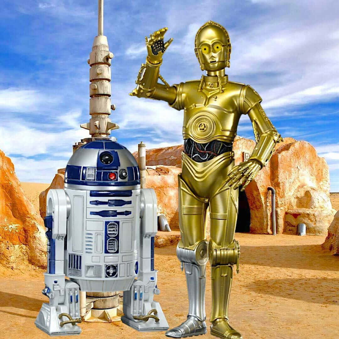 R2-D2與C-3PO塔圖因背景 壁紙 — 兩個標誌性的星球大戰機械人R2-D2和C-3PO站在沙漠峽谷中，頭頂是明亮的藍天，散佈著幾朵雲。