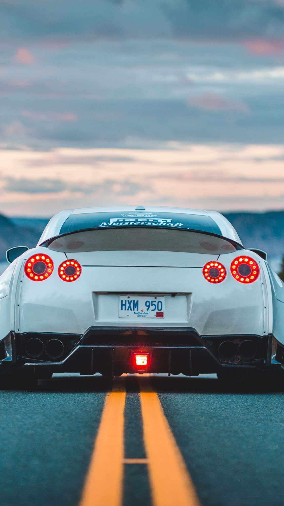 路中央的白色R33 GT-R 壁紙 — 一輛白色跑車亮著紅色尾燈，在黃昏時分行駛在道路上，背景是山脈。