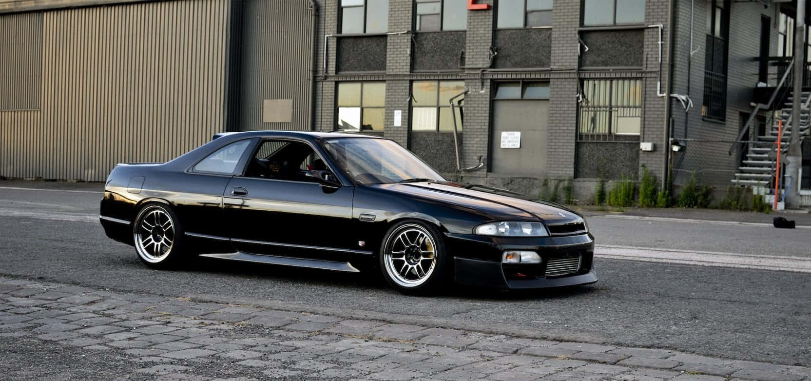 R33 GTR駛過城市街道 壁紙 — 一架時尚的黑色日產Skyline雙門跑車停泊在工業建築旁的鵝卵石街道上。