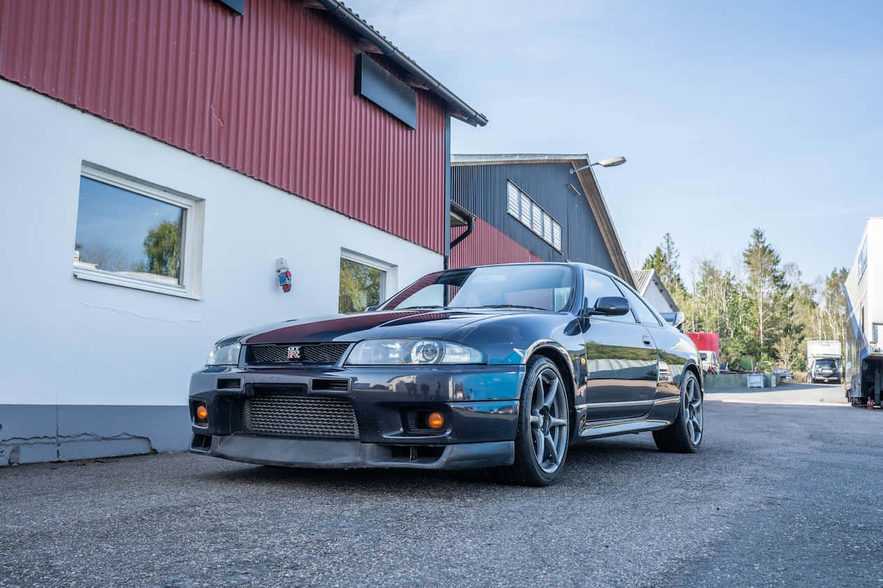 R33 GTR性能超跑疾馳向前 壁紙 — 一架時尚的黑色日產Skyline GT-R停喺紅白相間嘅工業大廈前面，藍天白雲。