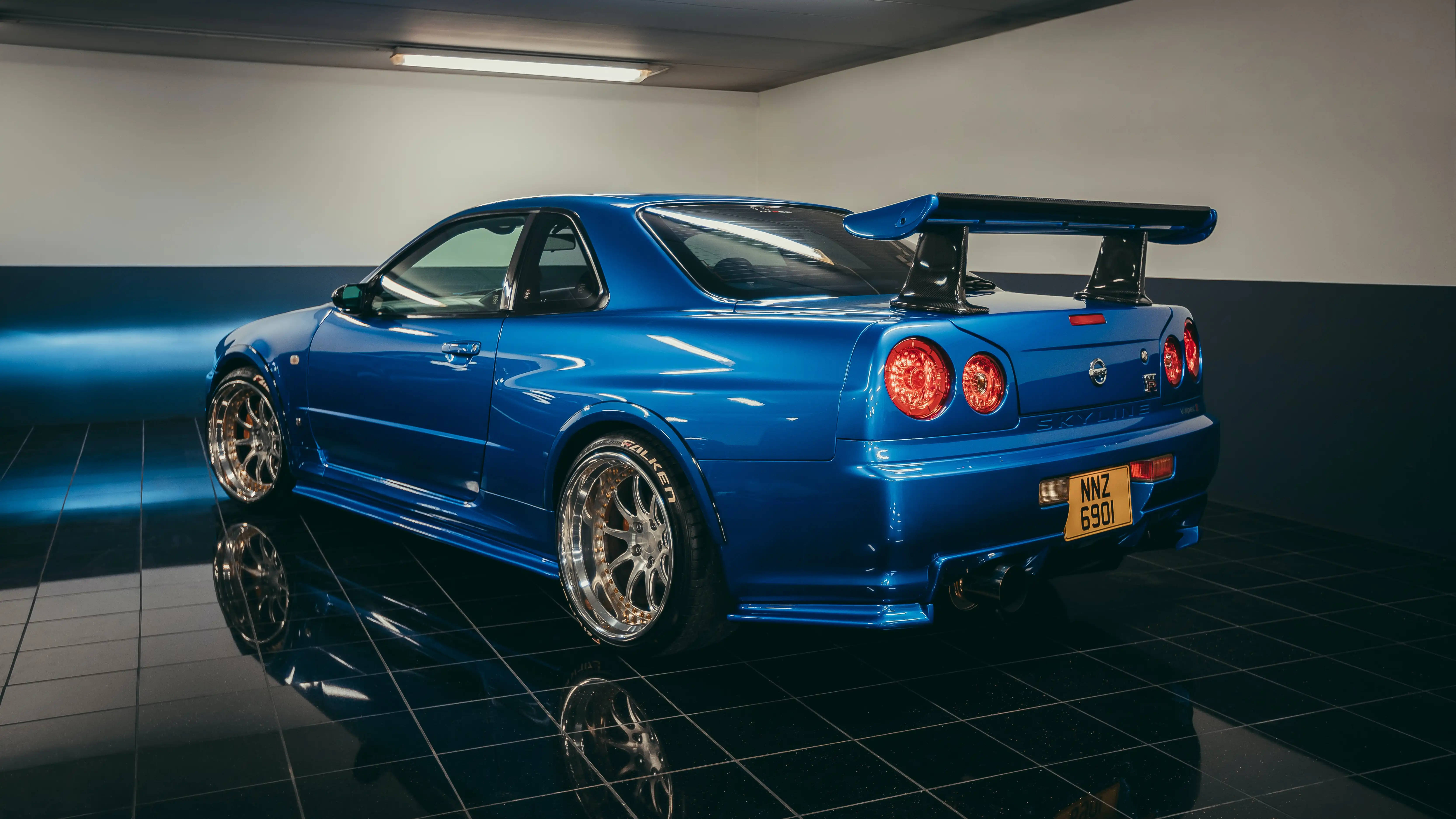 經典藍色 R34 GT-R 車款 壁紙 — 一輛光澤藍色的日產Skyline R34跑車，帶有大型後擾流板，在光線昏暗、舖有反光黑色瓷磚地板的車庫中展示。
