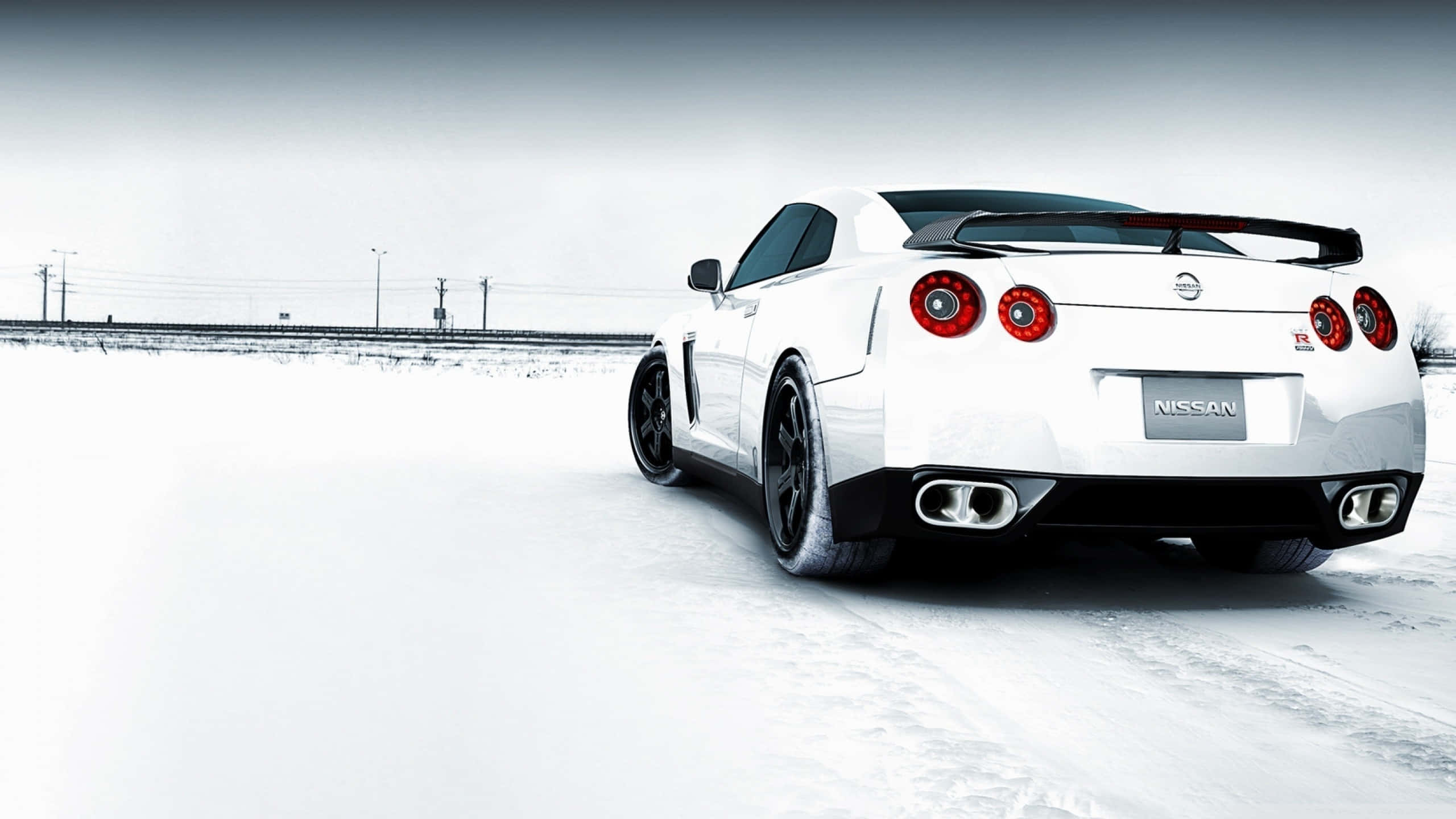尼桑R35 GT-R雪地道路 壁紙 — 一輛白色日產GT-R跑車，紅色尾燈，在灰色天空下駛過雪地。