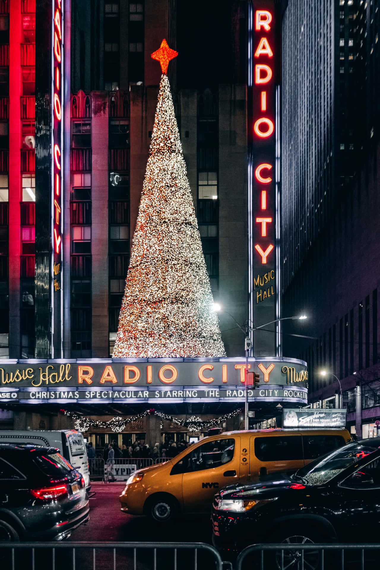 Radio City FM 簡約可愛聖誕燈樹 壁紙 — 一棵高聳的聖誕樹閃爍著燈光，自豪地矗立在標誌性的無線電城音樂廳前，夜晚被高聳的建築物和黃色的士環繞。