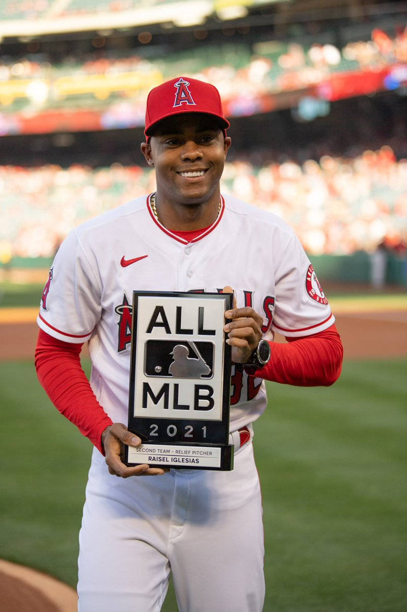 Raisel Iglesias - 一個驕傲的姿勢 壁紙 — 一位微笑的棒球運動員身穿白色和紅色的天使隊制服，在陽光明媚的球場上手持 All MLB 2021 獎項，身後是模糊的體育場人群。