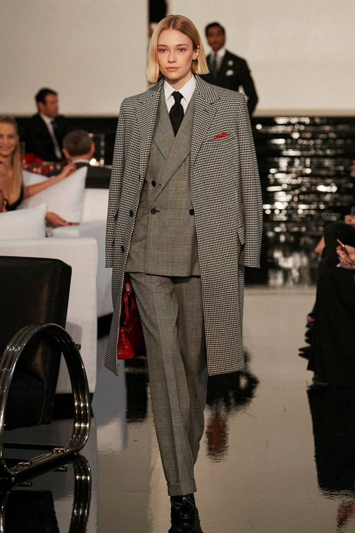 Ralph Lauren 女士西裝造型 壁紙 — 一位模特兒自信地走在閃亮的T台上，身穿千鳥格大衣和配套西裝，手持紅色手袋。