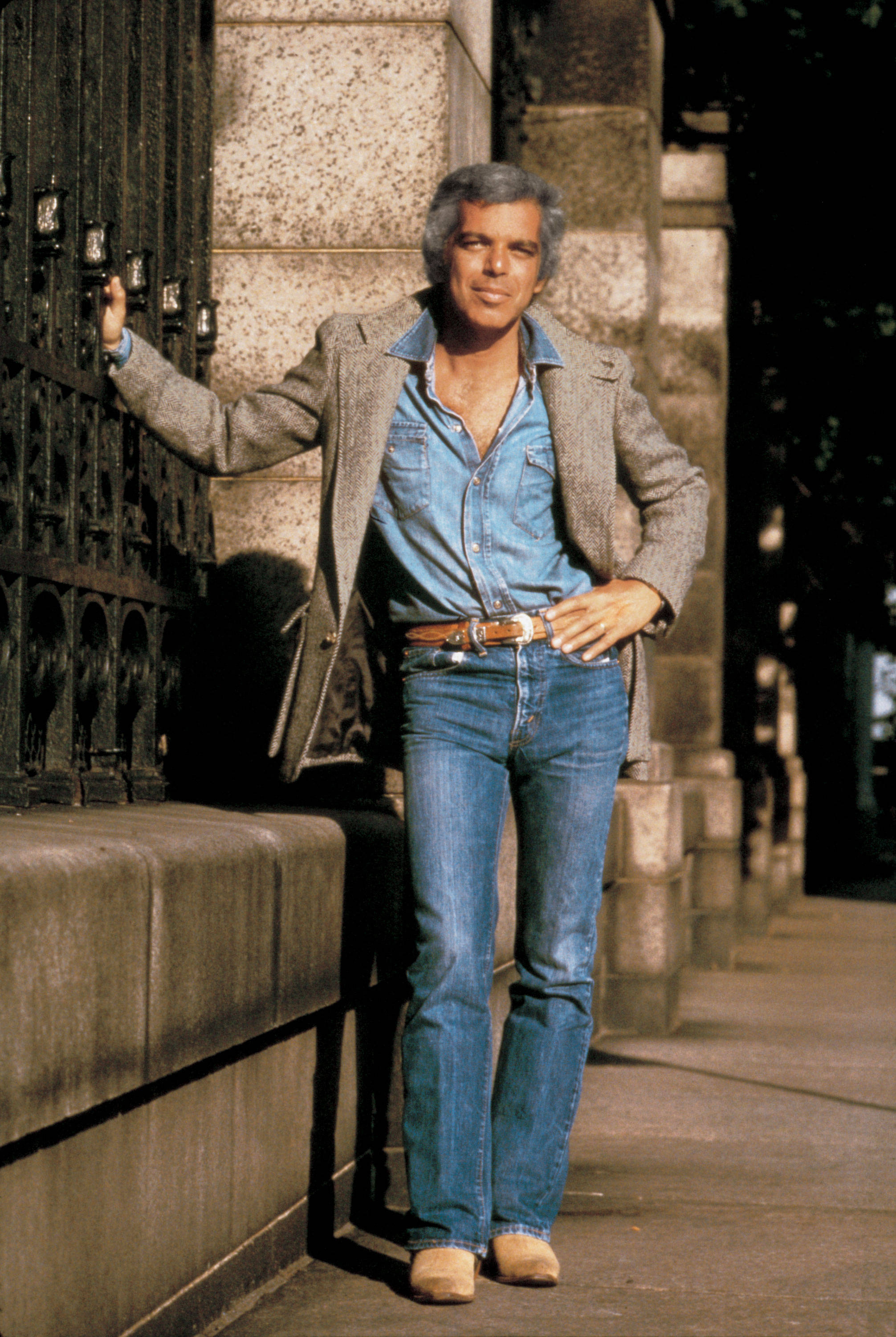 Ralph Lauren 巴黎 壁紙 — 一位穿著牛仔布和西裝外套的時尚男士，在陽光明媚的城市人行道上，隨意地靠在裝飾華麗的大門旁的石牆上。