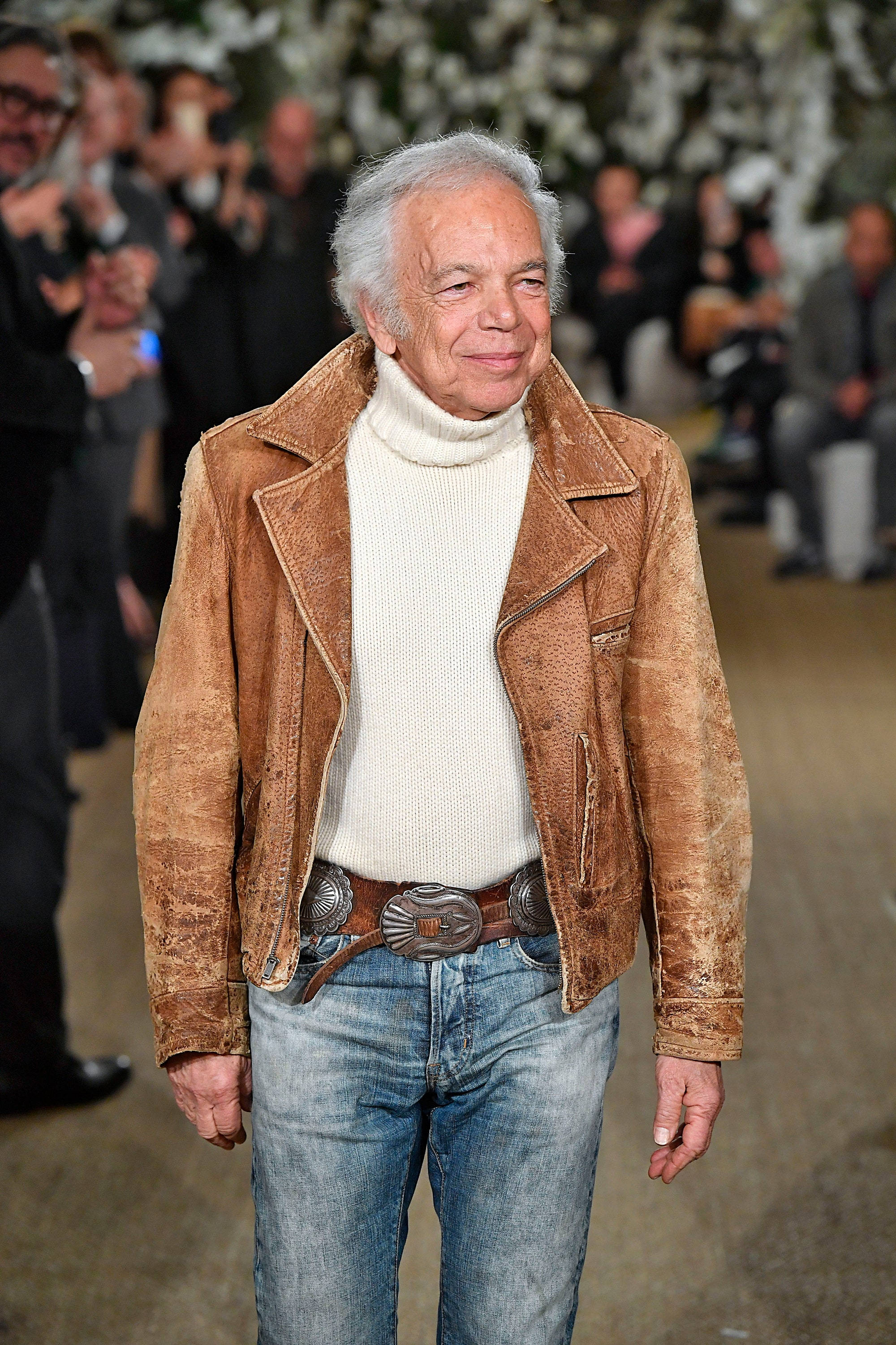 Ralph Lauren 毛衣 壁紙 — 一位白髮年長男士穿著棕色麂皮外套內搭白色高領毛衣和牛仔褲微笑。