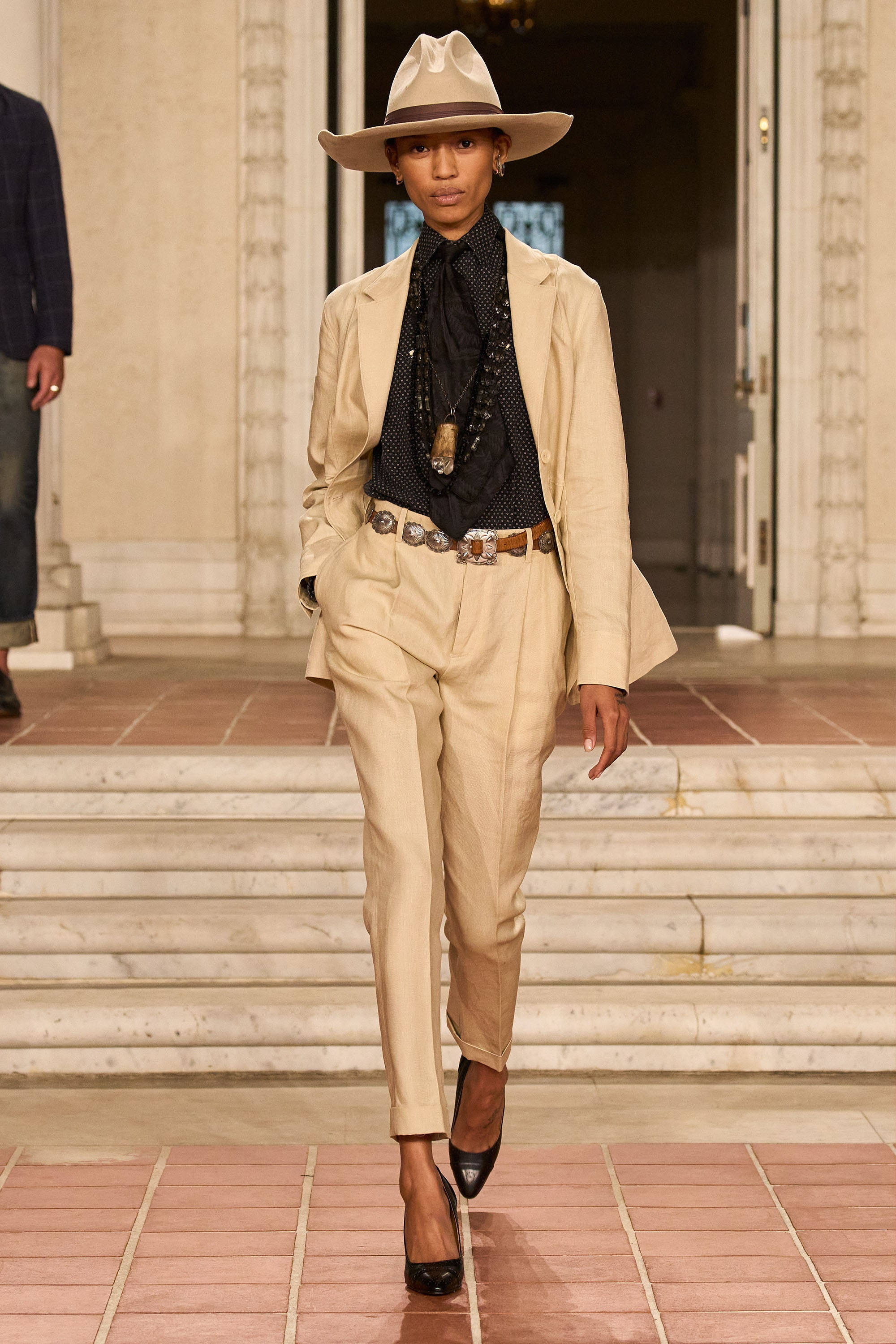 Ralph Lauren 非洲探險風 壁紙 — 一位模特兒自信地走在瓷磚跑道上，身穿米色西裝、牛仔帽和黑色高跟鞋。