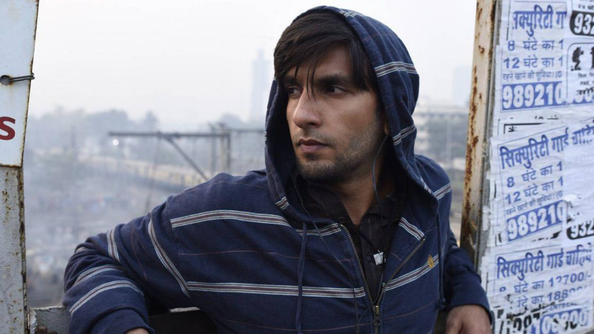 Ranveer Singh 在《Gully Boy》電影中 壁紙 — 一個穿著藍色條紋連帽衫的年輕人，在生鏽的月台上若有所思地凝視著，火車穿過朦朧的城市背景。