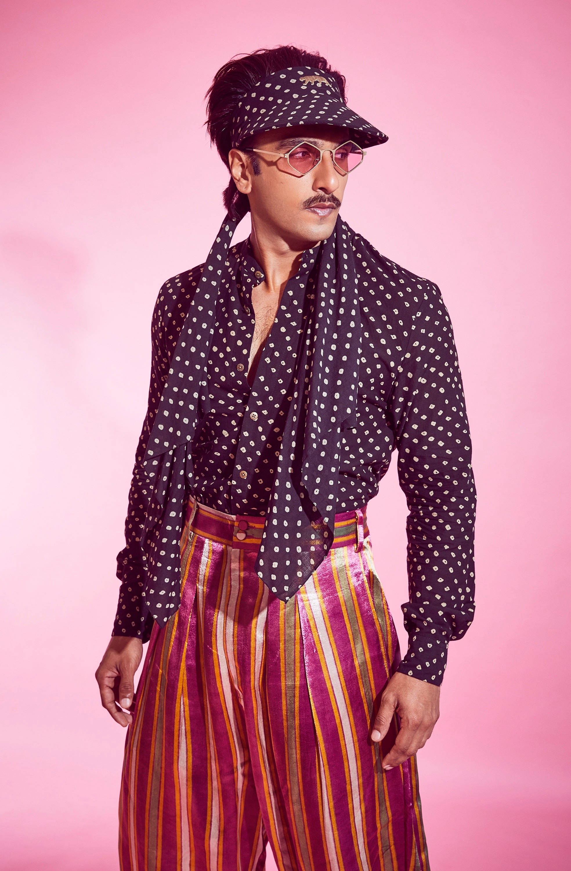 Ranveer Singh 的時尚奇裝異服 壁紙 — 一位時尚男士穿著波點襯衫和條紋褲，在純粉紅色背景前自信地擺姿勢。