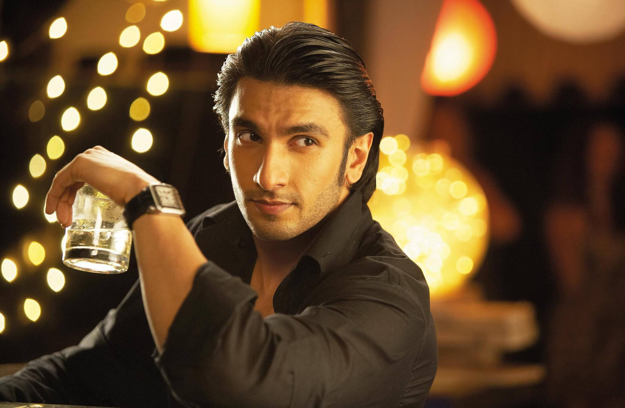 Ranveer Singh 在《Ladies vs Ricky Bahl》電影中 壁紙 — 一個穿著黑色襯衫的男人在一個溫暖照明的酒吧裡，背景有串燈和發光球體，手持一杯水。