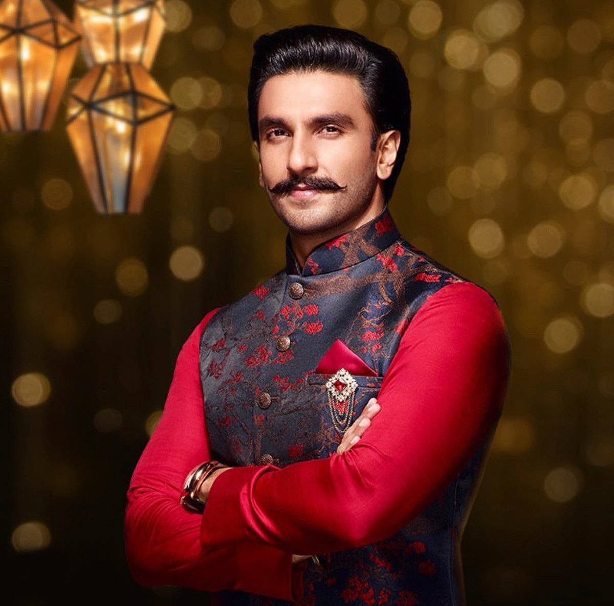Ranveer Singh 為 Manyavar 品牌拍攝代言照 壁紙 — 一個留鬍子的男人，身穿紅色圖案背心，雙臂交叉，自信地站在金色散景背景前，背景有懸掛的燈籠。