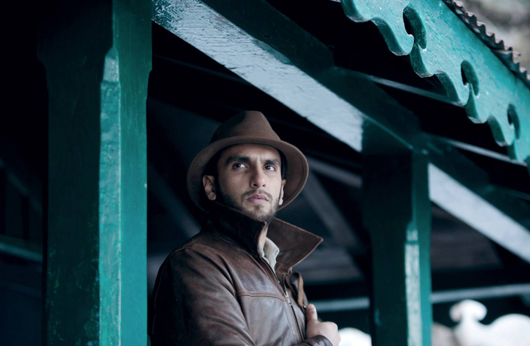 Ranveer Singh 飾演 Varun Srivastav 壁紙 — 一個戴著棕色軟呢帽、身穿皮褸的男人，在飽經風霜的綠色木簷下若有所思地凝視。