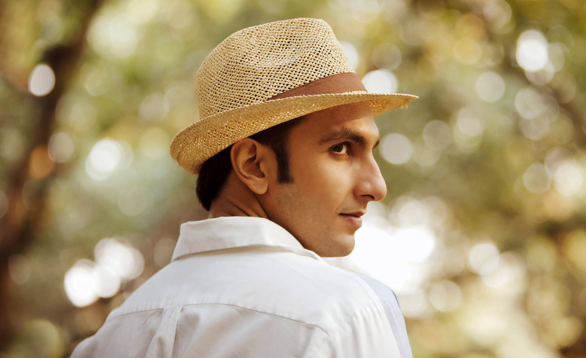 Ranveer Singh 在電影《Lootera》中的造型 壁紙 — 一個戴草帽、穿白恤衫的男人，在陽光普照的散景森林背景下，轉頭越過肩膀向後看。