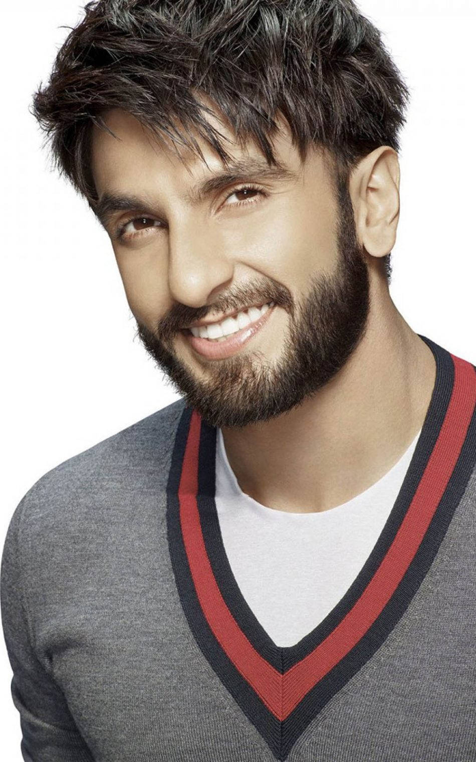 Ranveer Singh 微笑特寫 壁紙 — 一位微笑的黑髮鬍鬚男士，身穿灰色V領毛衣，帶有紅色和黑色飾邊，背景為白色。