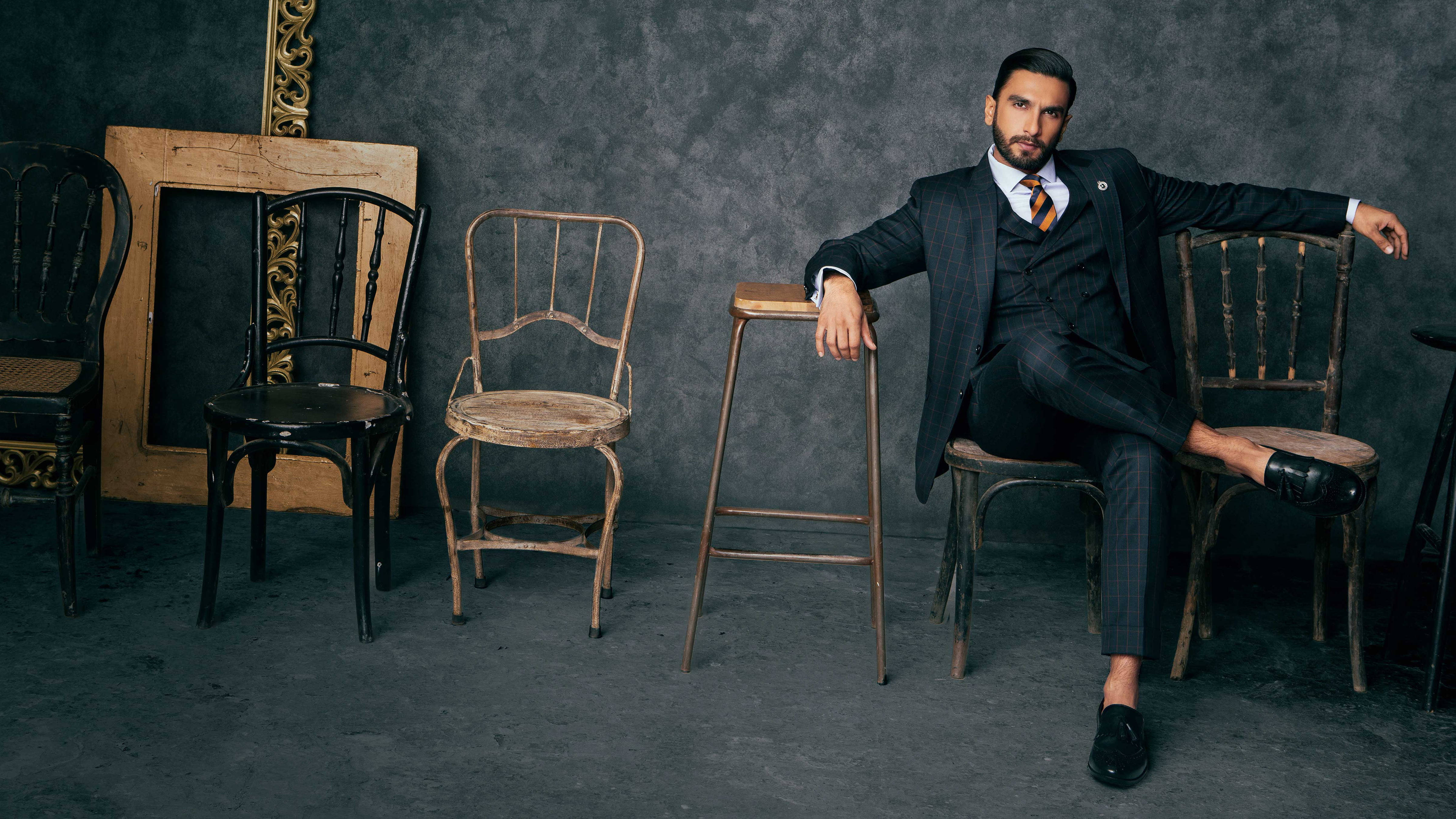 Ranveer Singh 為 Maxim India 拍攝寫真 壁紙 — 一位穿著深色西裝、衣著講究的男子，在質感灰色牆壁前，在一堆復古椅子中的一張鄉村風格椅子上自信地閒坐。