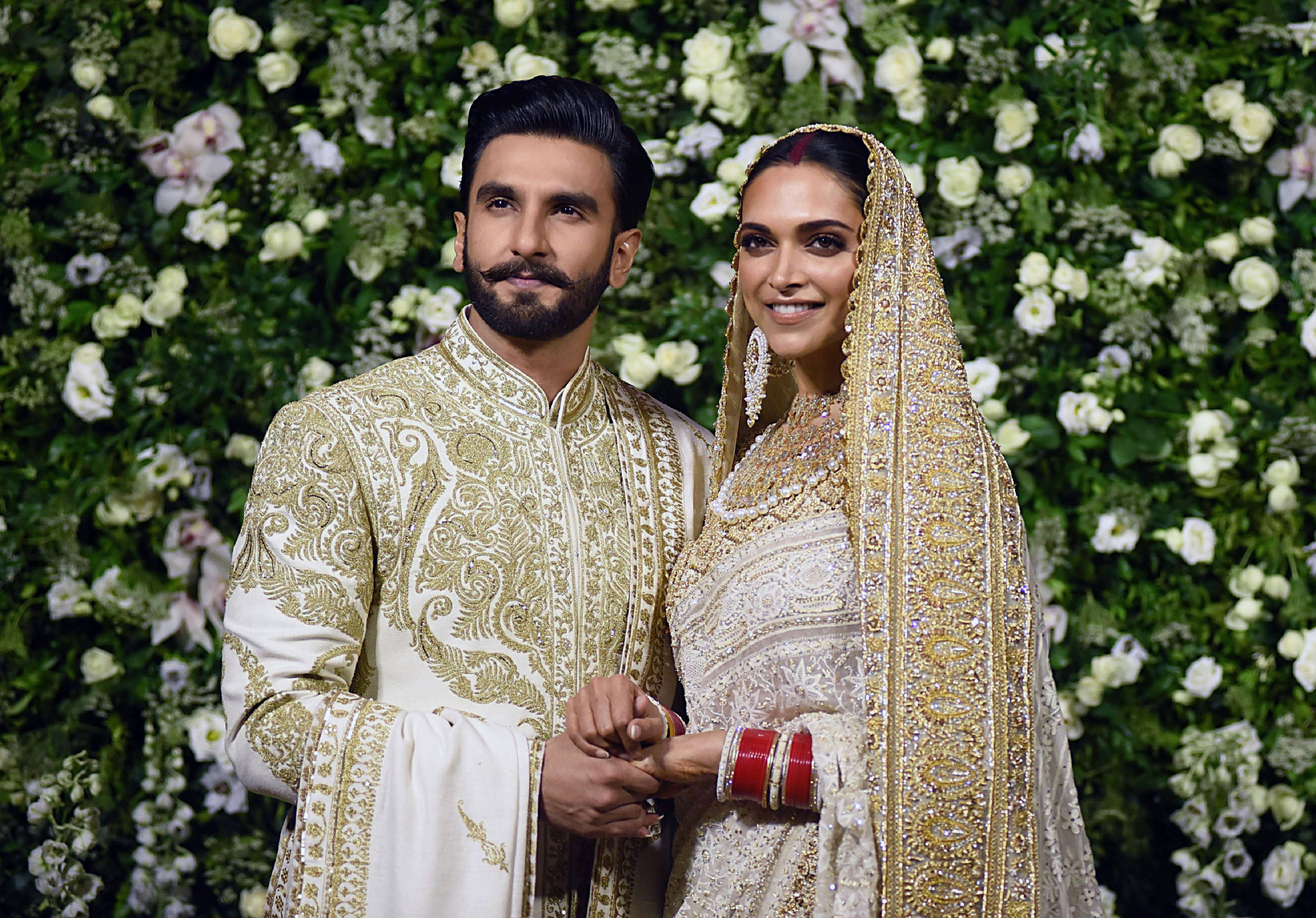 Ranveer Singh 與 Deepika Padukone 的婚禮盛況 壁紙 — 一對閃耀的新人穿著華麗的白色和金色婚禮服飾，在茂盛的花卉背景前牽手。