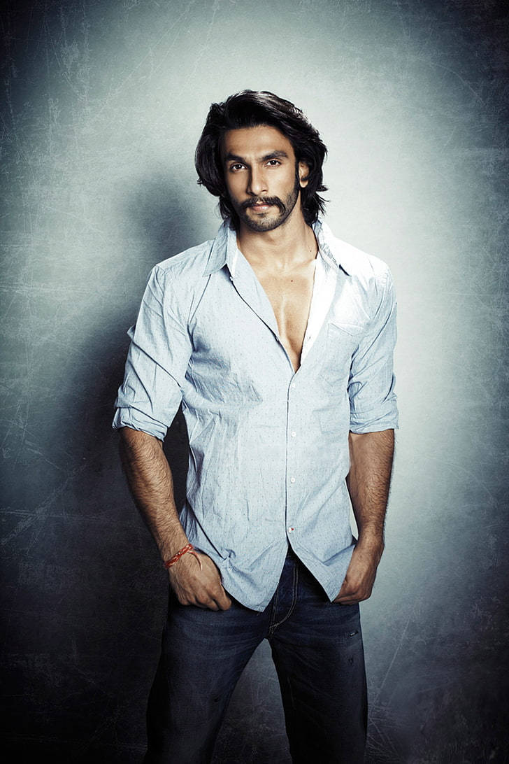 Ranveer Singh 飾演 Bikram 壁紙 — 一位自信的黑髮鬍鬚男，穿著淺藍色開胸襯衫和牛仔褲，背景是質感灰色攝影棚。