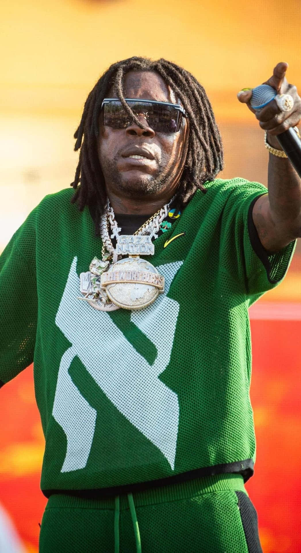  壁紙 — 一位戴著辮子頭和太陽眼鏡的饒舌歌手在舞台上表演，身穿綠色網眼毛衣，上面有大型白色標誌和多條金鏈。