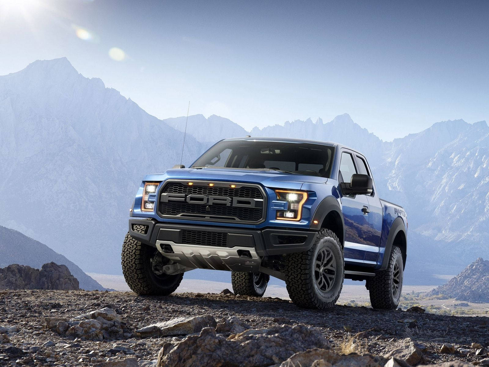 福特Raptor越野行駛 壁紙 — 一輛藍色福特F-150 Raptor卡車停在岩石地形上，背後有白雪覆蓋的山脈在明亮的陽光下聳立。