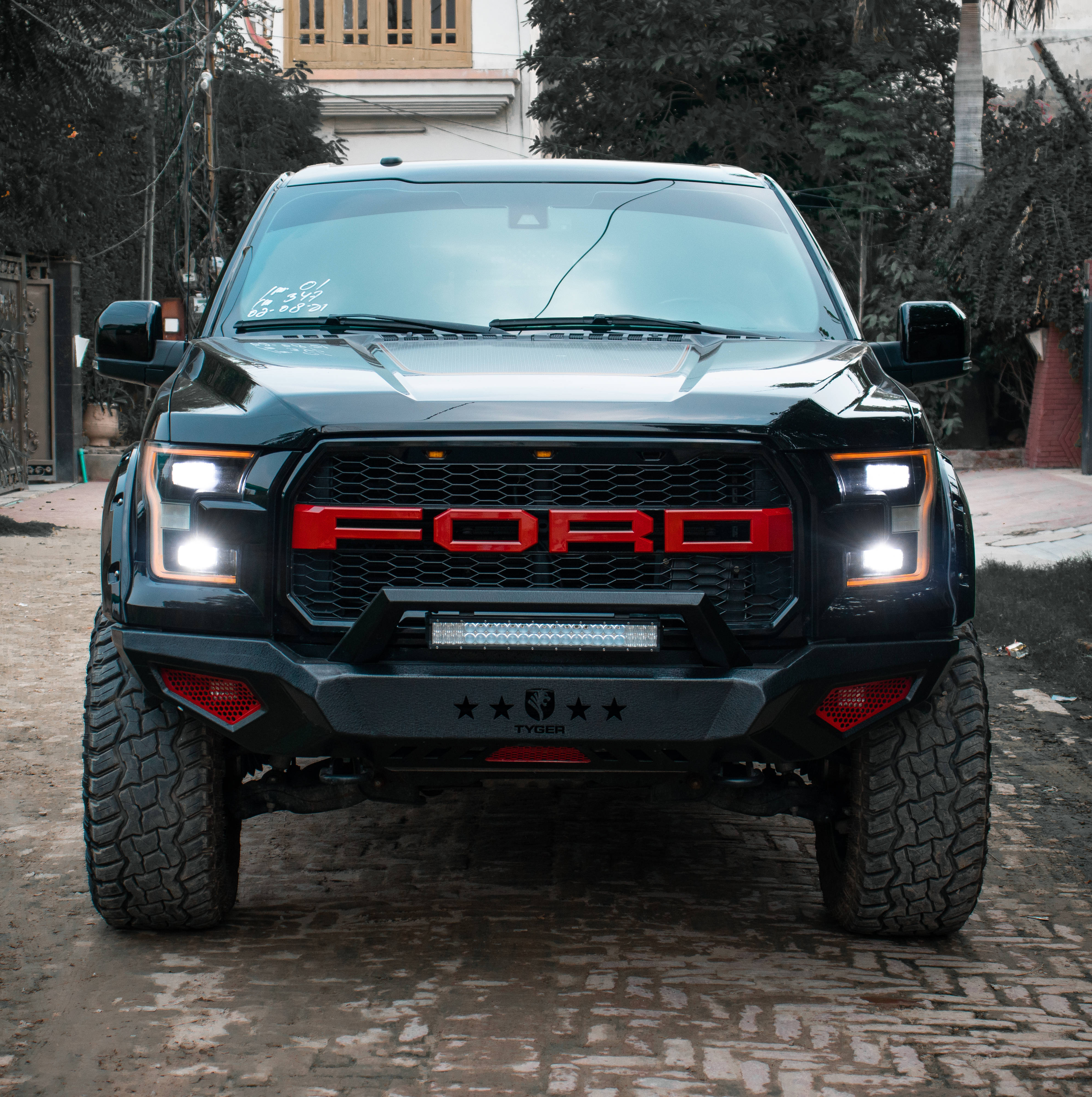 光澤黑漆福特Raptor 壁紙 — 一輛黑色福特F-150 Raptor帶有紅色點綴和發光車頭燈，停在鵝卵石街道上，周圍環繞著樹木和建築物。