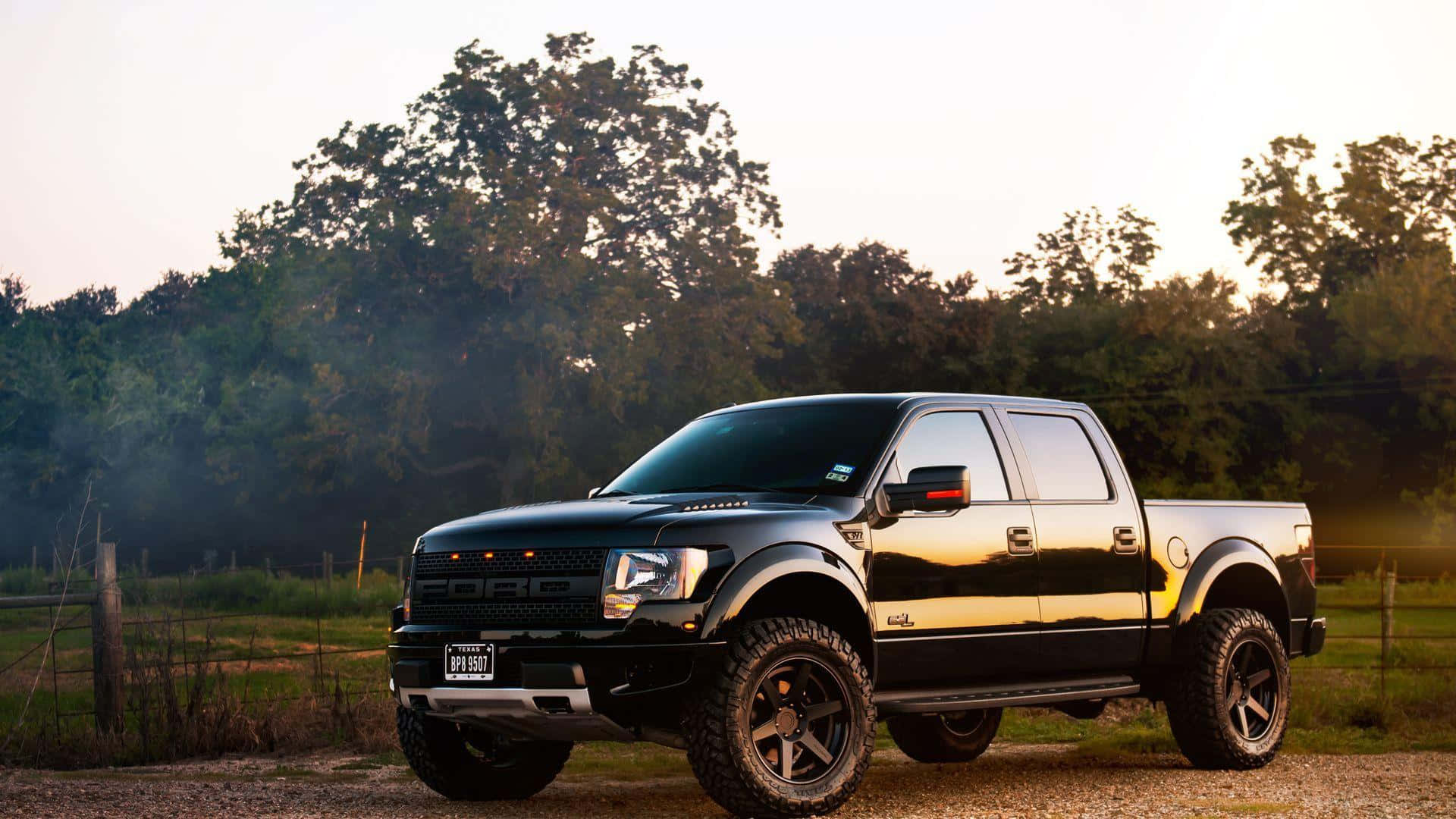 黑色福特 Raptor 舉高貨車 壁紙 — 一輛黑色福特F-150皮卡車配備大型輪胎，在日落時分停在碎石路上，周圍環繞著樹木和木柵欄。