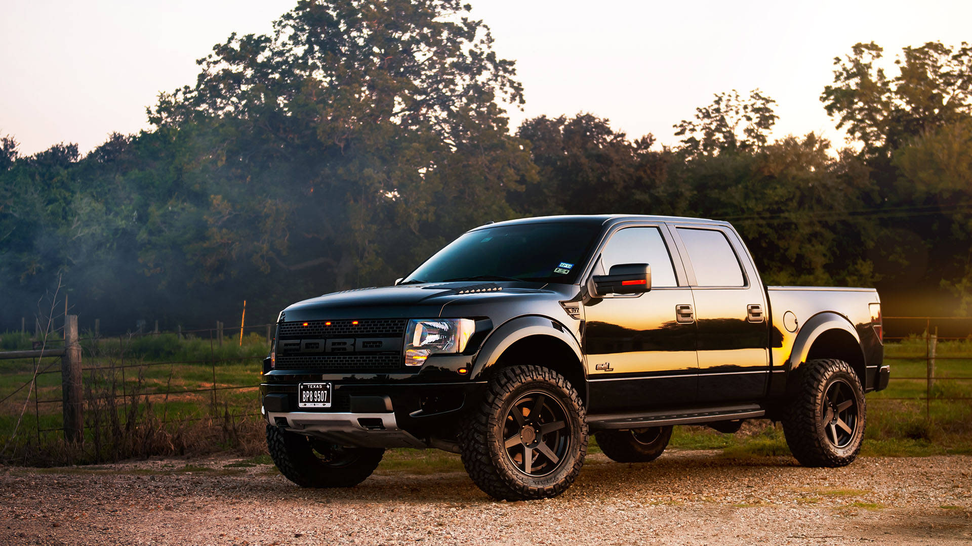 光澤黑福特 Raptor 頂級風采 壁紙 — 一輛黑色福特F-150皮卡車，配備升高懸掛和大輪胎，在日落時分停在碎石路上，周圍有樹木和木柵欄。