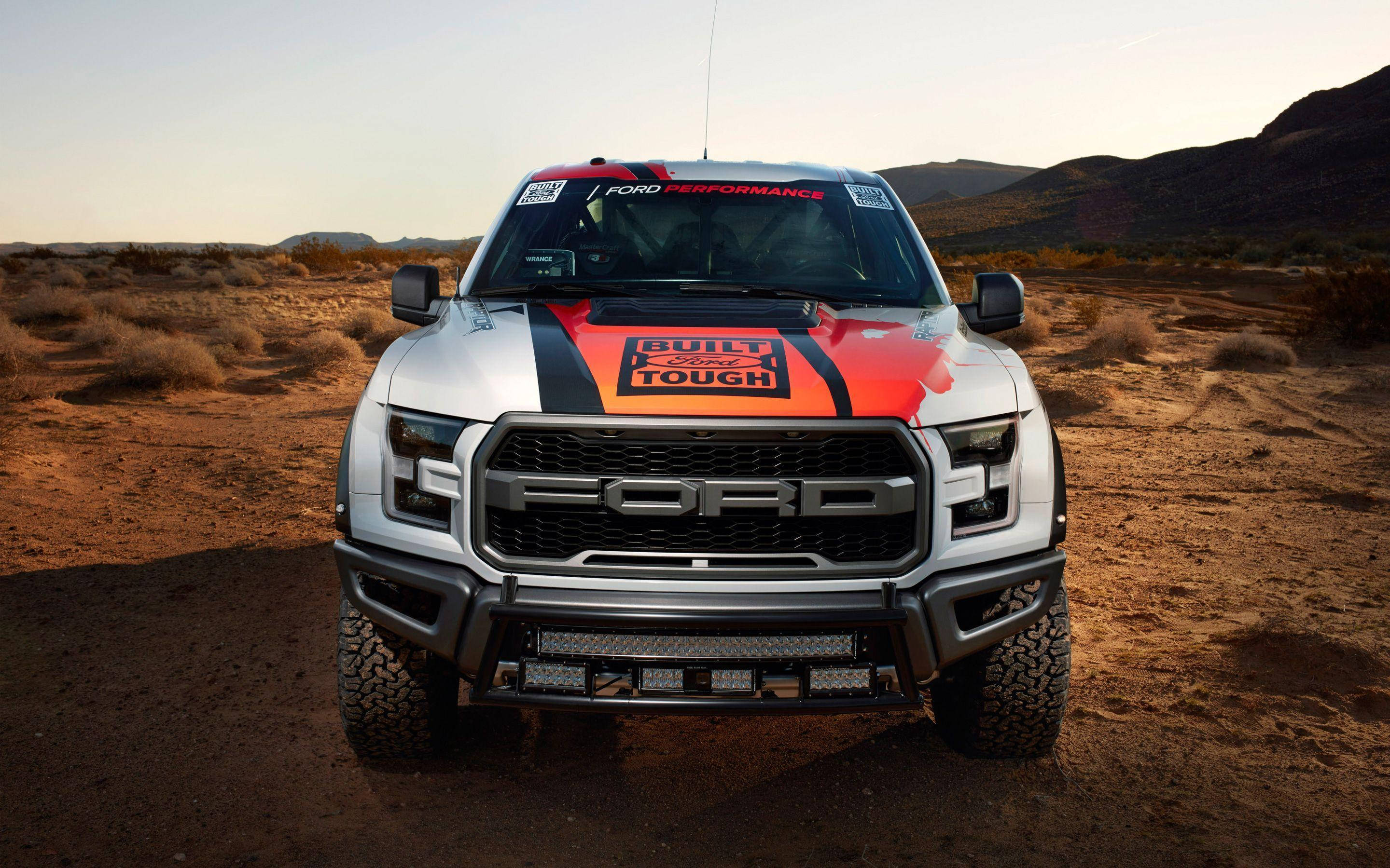 自訂白色塗裝福特Raptor 壁紙 — 一輛銀色福特F-150 Raptor帶有橙色和黑色賽車貼花，在陽光明媚的沙漠景觀中傲然矗立。