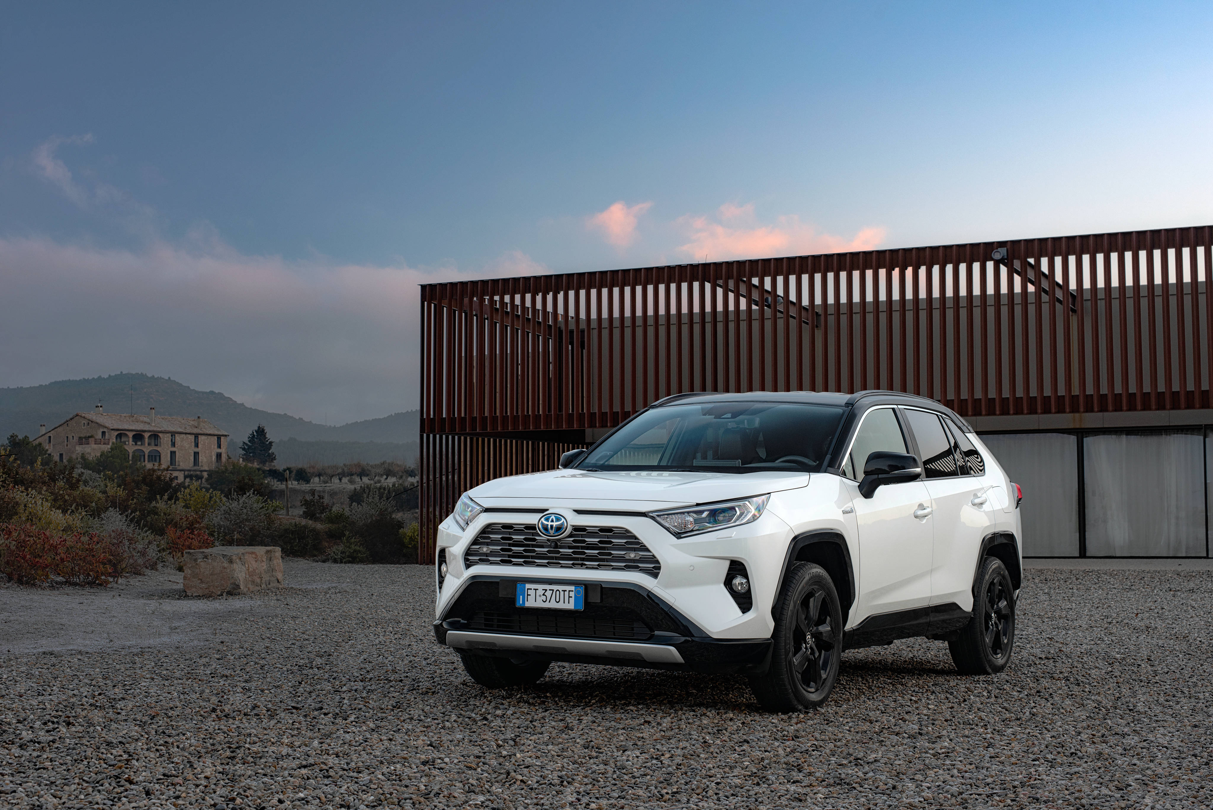 白色豐田 RAV4 4K 壁紙 — 一輛白色豐田RAV4 SUV停泊在碎石上，旁邊是現代木柵欄，背景有山脈和石建築，在黃昏時分。