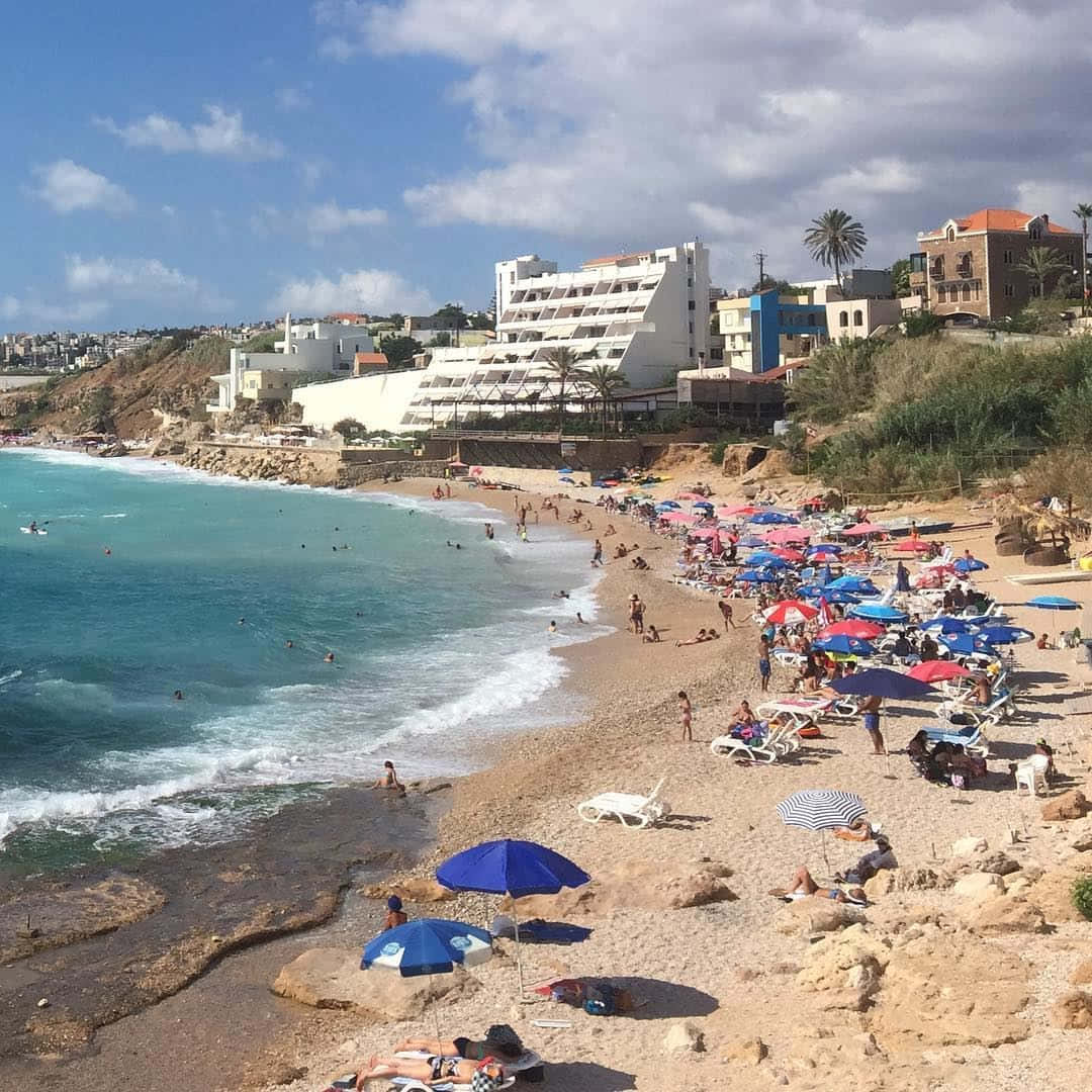 Mediterranean Beach 桌布 壁紙 — 一個熱鬧的海灘場景，碧綠色的海浪拍打著岩石海岸，岸邊排列著色彩繽紛的遮陽傘和享受日光浴的人們，天空湛藍明亮。