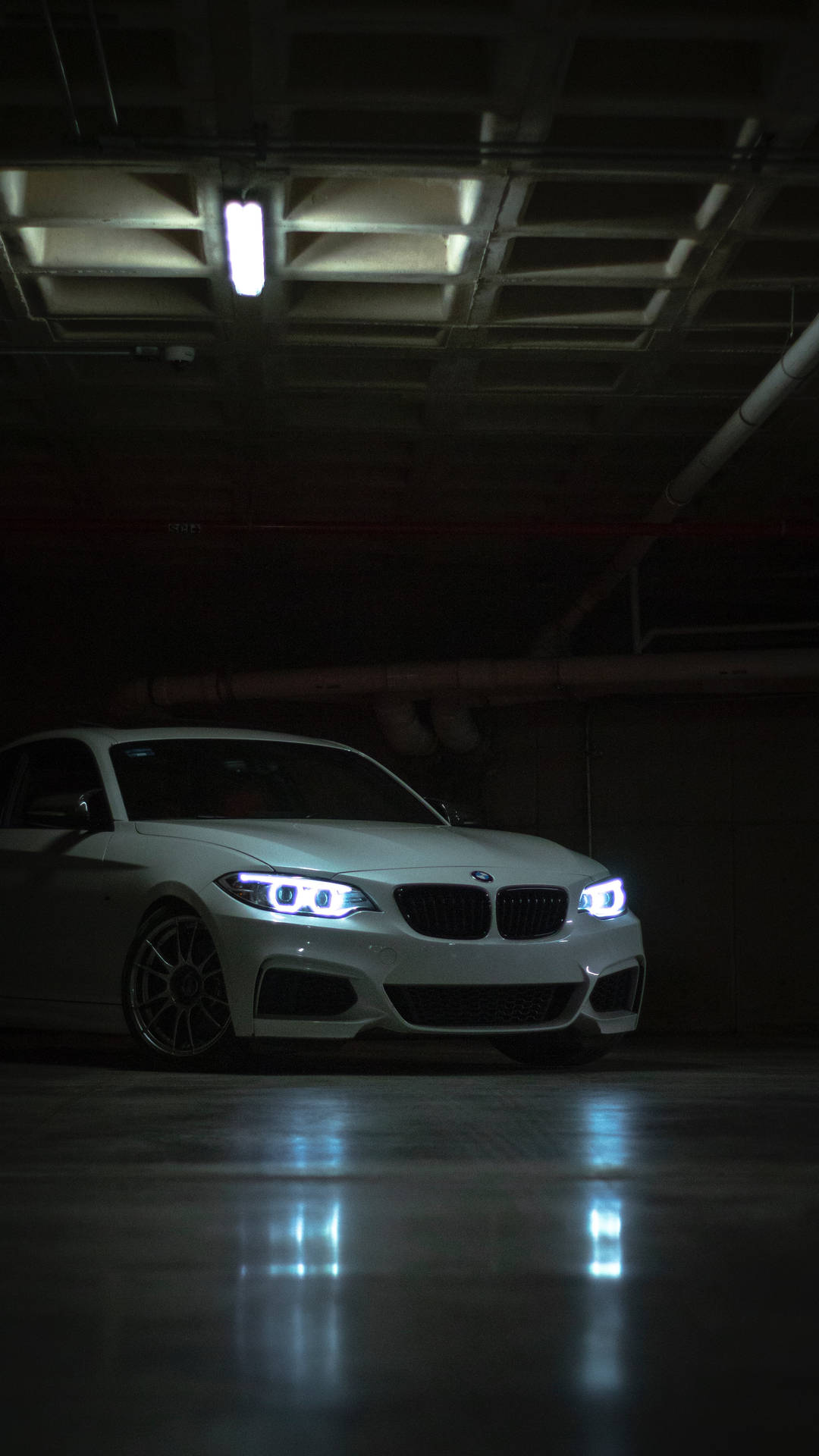 白色BMW汽車，車頭燈明亮，停泊在黑暗的地下停車場，地板濕滑有倒影。