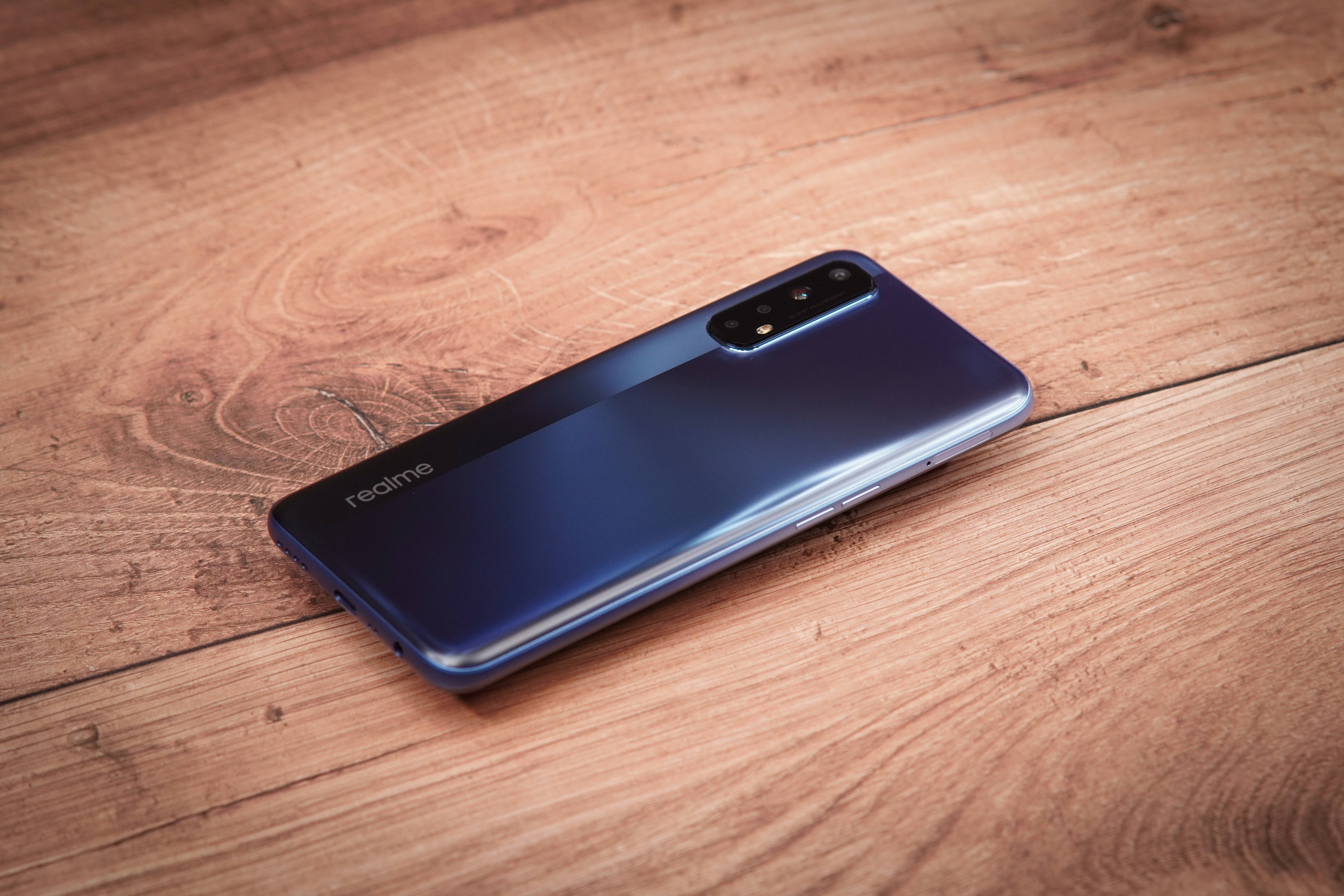 Realme 7 黑色款 壁紙 — 一部時尚的藍色 realme 智能手機對角放置在溫暖的木質表面上，木紋清晰可見。