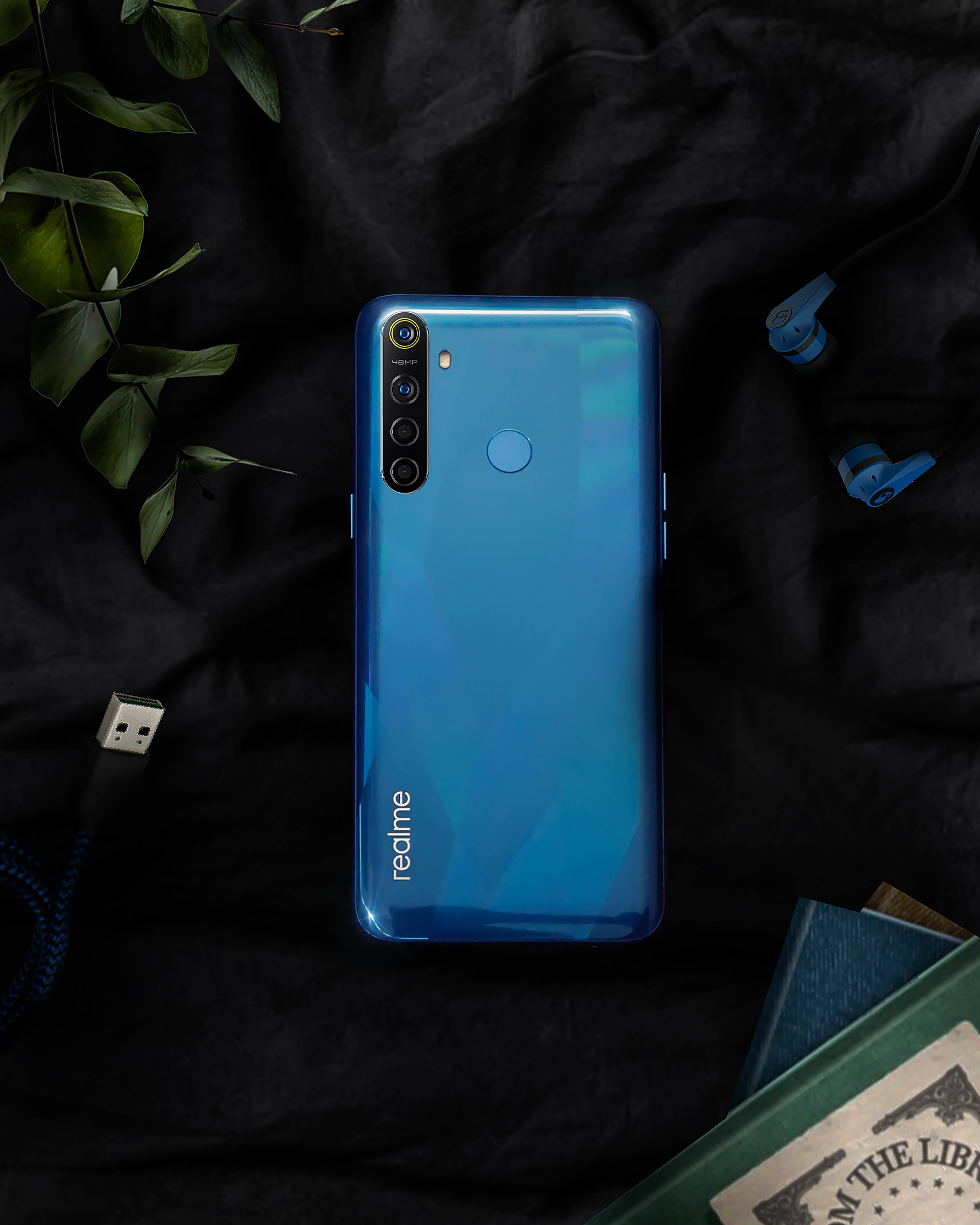 Realme 8手機 壁紙 — 一部亮藍色 realme 智能手機放在黑色布料上，周圍有綠色樹葉、耳塞和一本書。