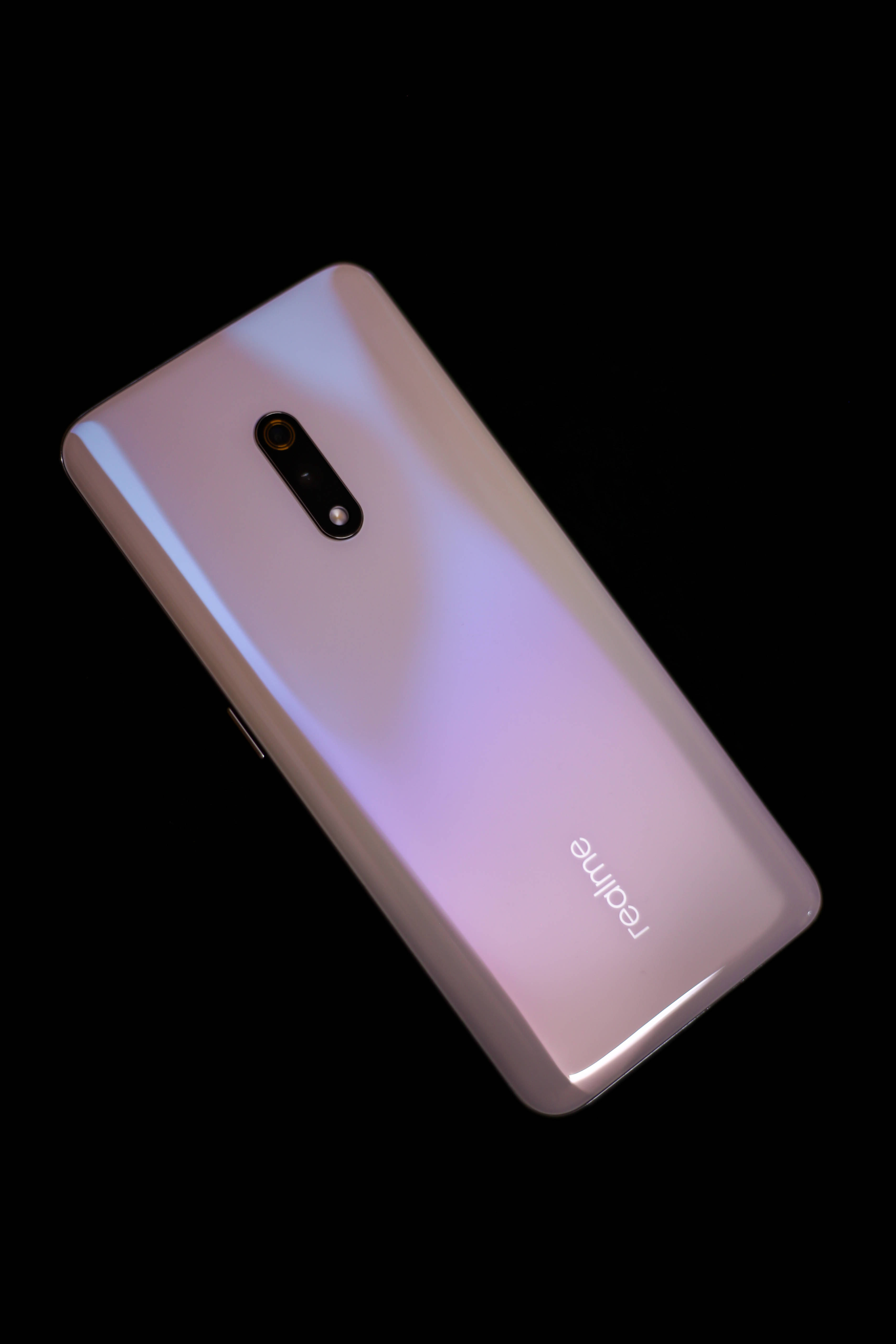 Realme X 粉色款 壁紙 — 一部時尚的漸變粉紅色 realme 智能手機，帶有垂直相機模組，斜靠在純黑色背景上。