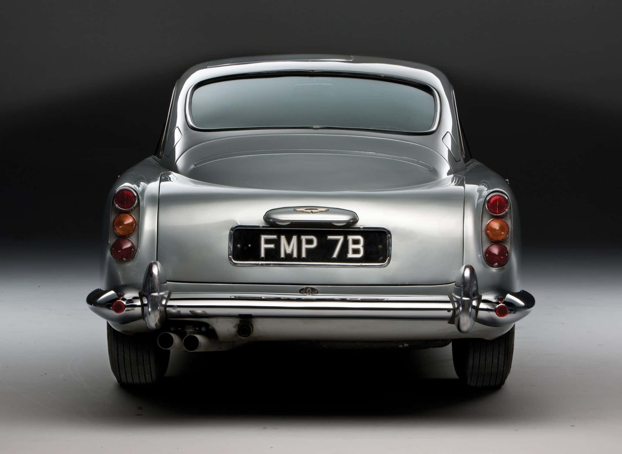 Aston Martin Db5 桌布 壁紙 — 從後方觀看一輛時尚的銀色Aston Martin DB5，帶有鍍鉻裝飾和紅色尾燈，背景為深色工作室。