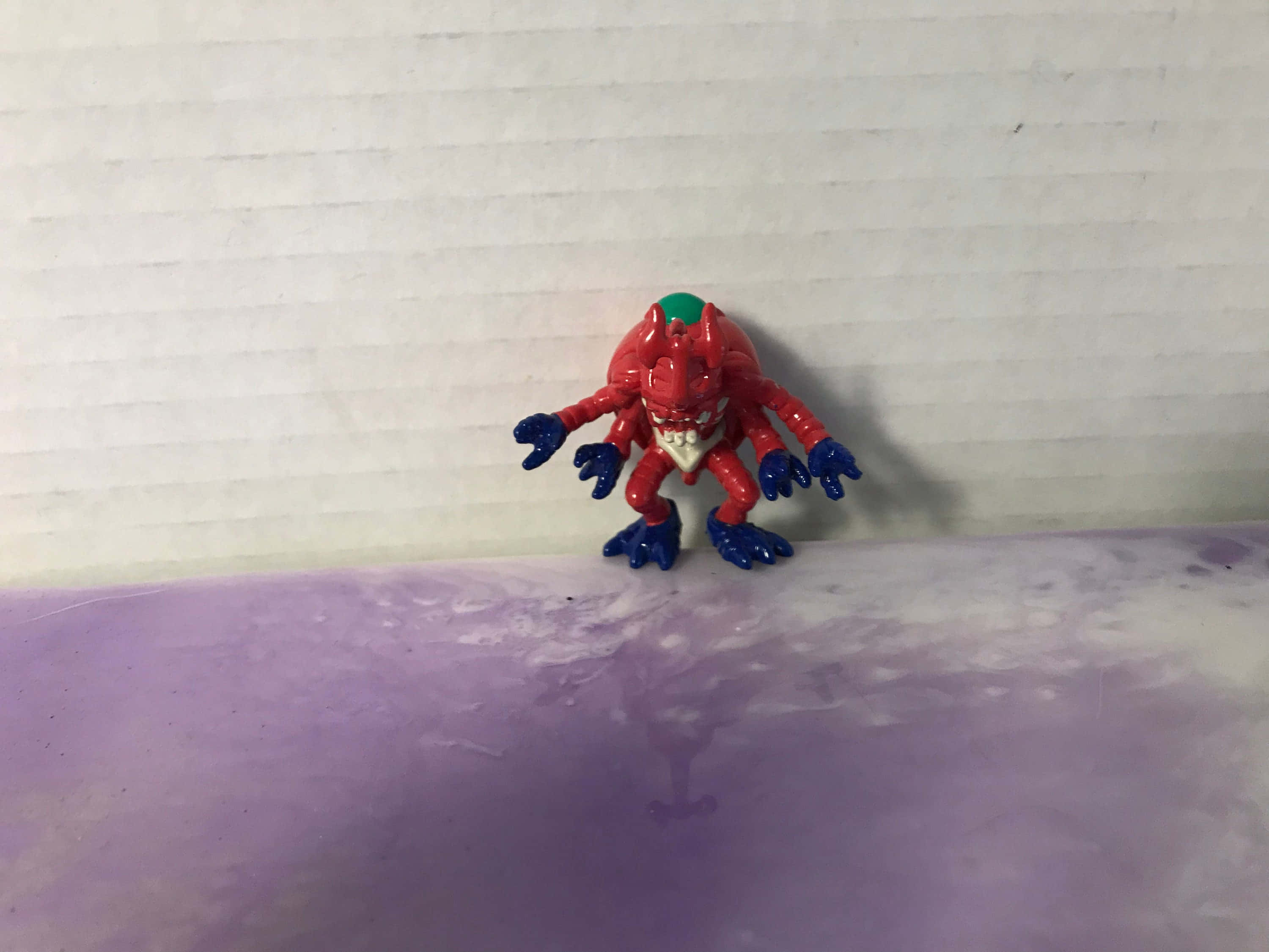Digimon Tentomon 桌布 壁紙 — 一個紅色和藍色的塑膠怪物玩具，有多條肢體，站在紫色表面上，背景是白色波紋牆。