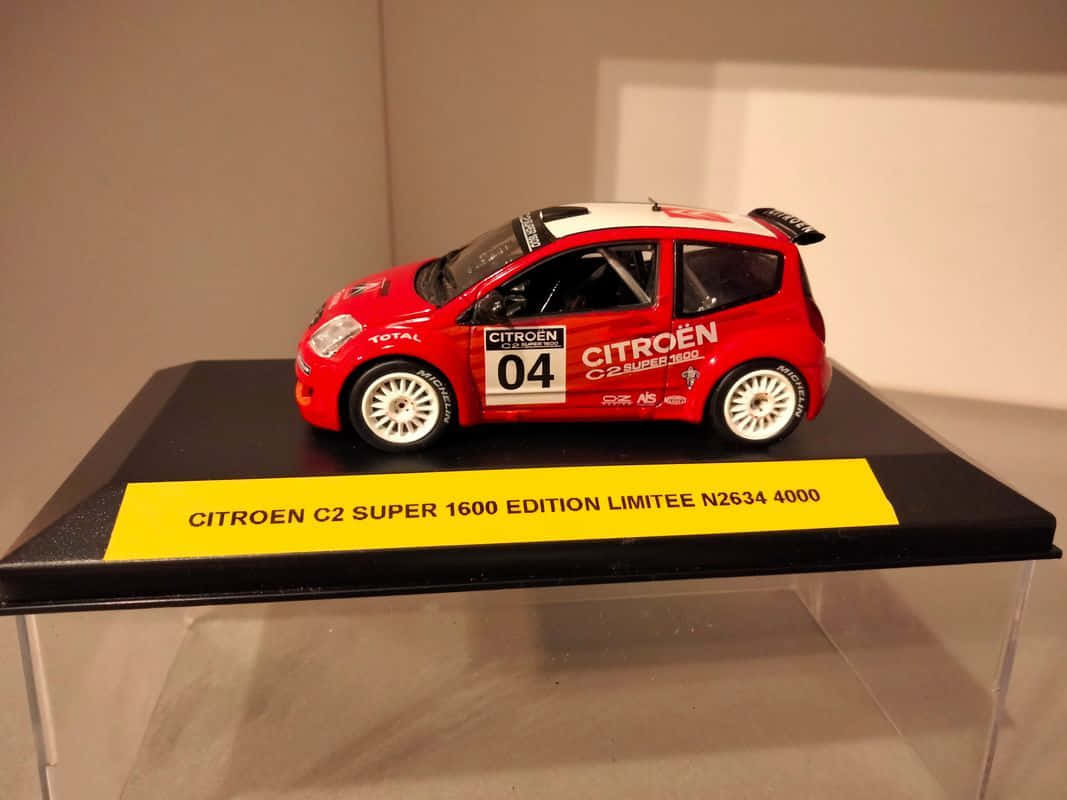 Citroen C2 Super 1600 桌布 壁紙 — 一輛紅白相間的雪鐵龍C2拉力賽車模型（編號04）放置在帶有黃色標籤的黑色展示架上，置於玻璃陳列櫃內。