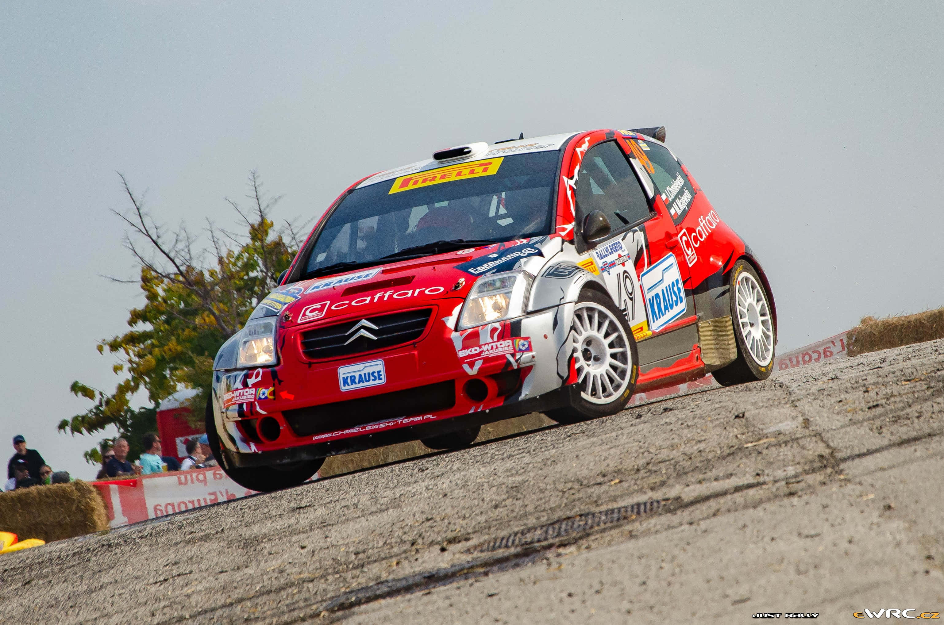 Citroen C2 Super 1600 桌布 壁紙 — 一輛紅白相間的拉力賽車在塵土飛揚的碎石賽道上飛馳，觀眾在乾草捆後觀看。