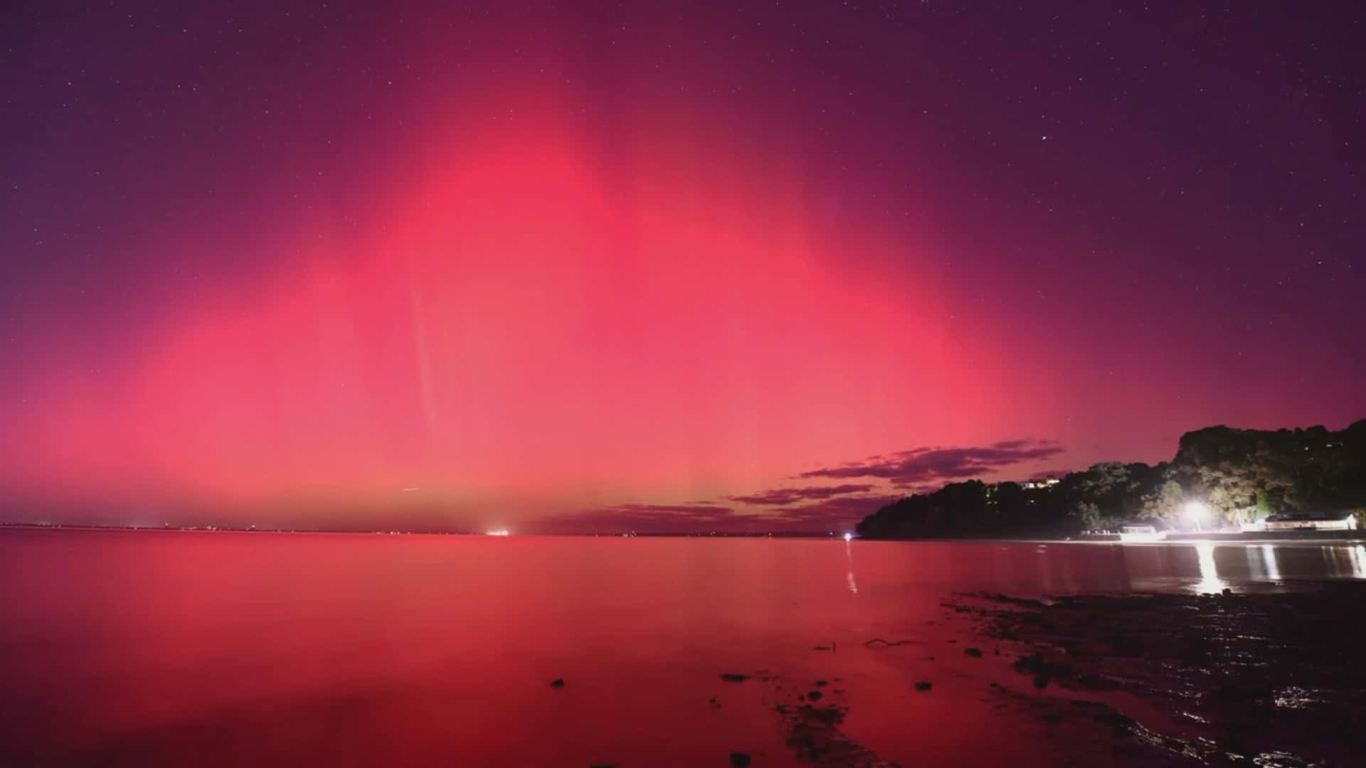  壁紙 — 鮮豔的紅色極光在繁星點點的夜空中舞動，映照在平靜的水面上，反射出遠處的燈光和黑暗的海岸線。