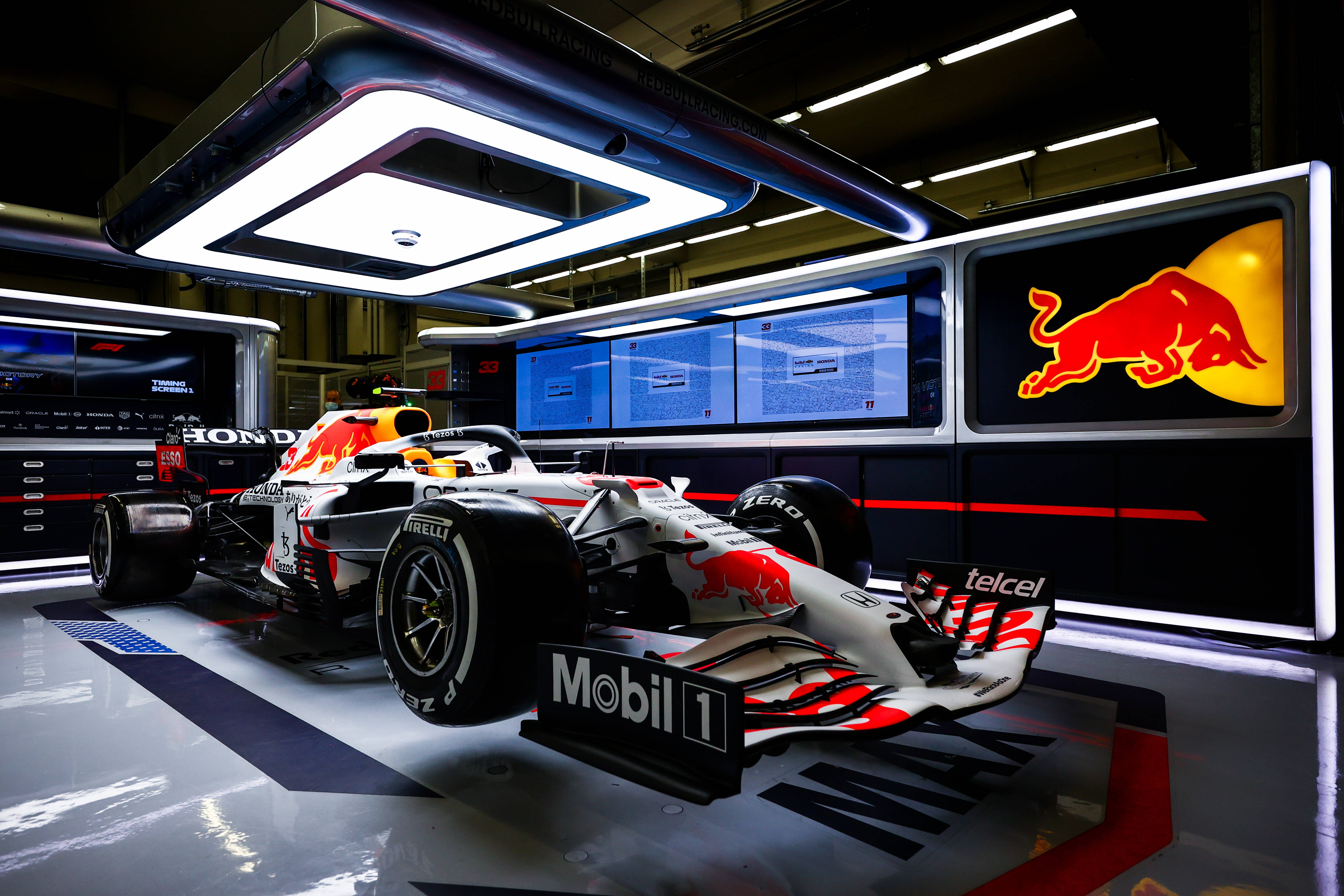 力量與速度的化身：Red Bull F1 賽車動態 壁紙 — 一部紅牛車隊一級方程式賽車在明亮的頂燈下閃閃發光，身處一個裝飾有贊助商標誌和數碼屏幕的高科技車庫。