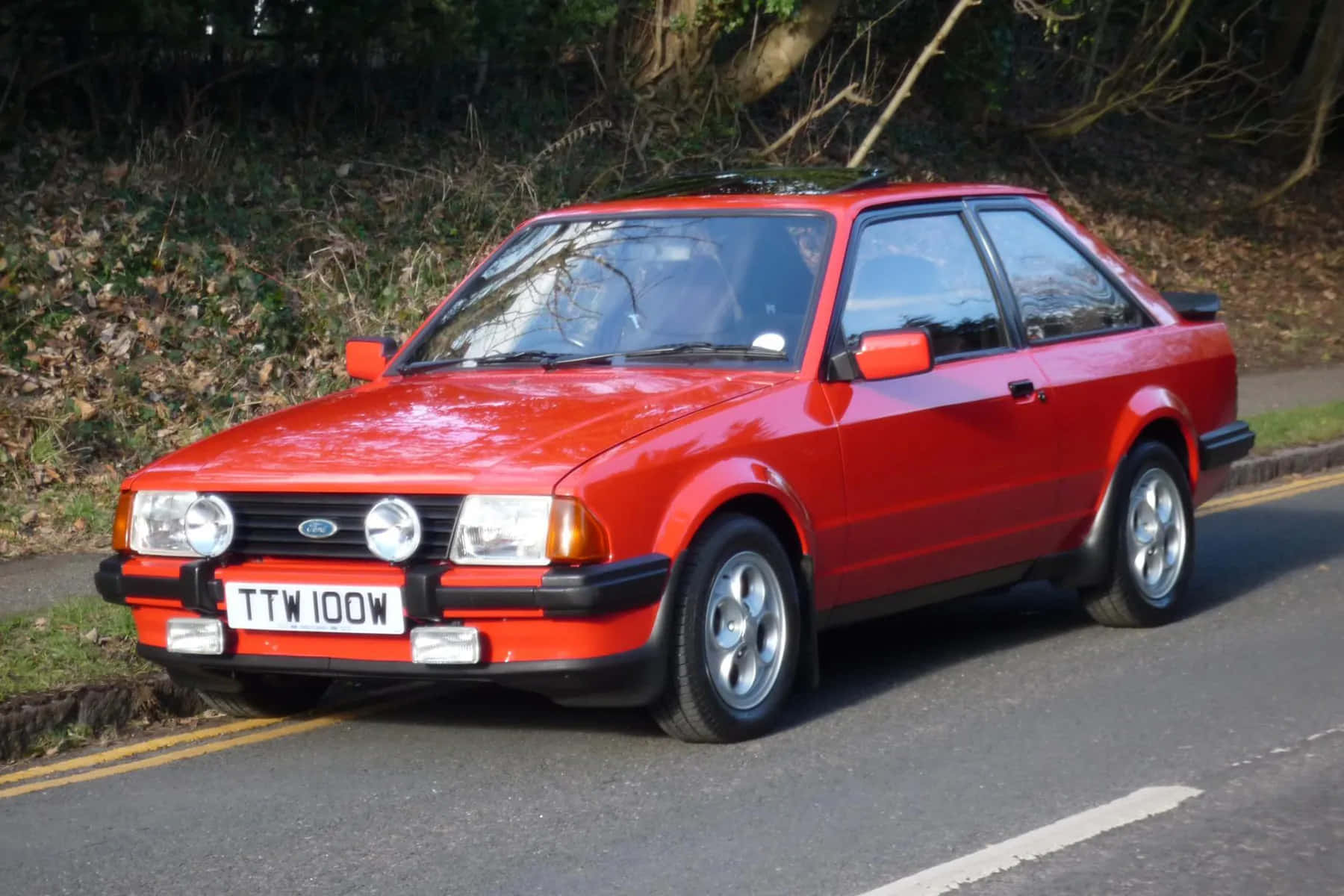 Ford Escort 桌布 壁紙 — 一輛鮮紅色的經典福特 Sierra Cosworth 停泊在路邊，背景是沒有葉子的樹木和草地。