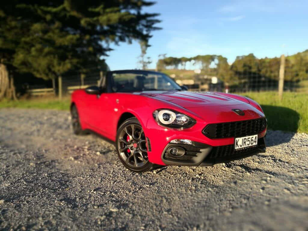Fiat 124 Spider 桌布 壁紙 — 一輛鮮紅色開篷跑車停泊在碎石路上，背景有樹木和籬笆，天空清澈湛藍。